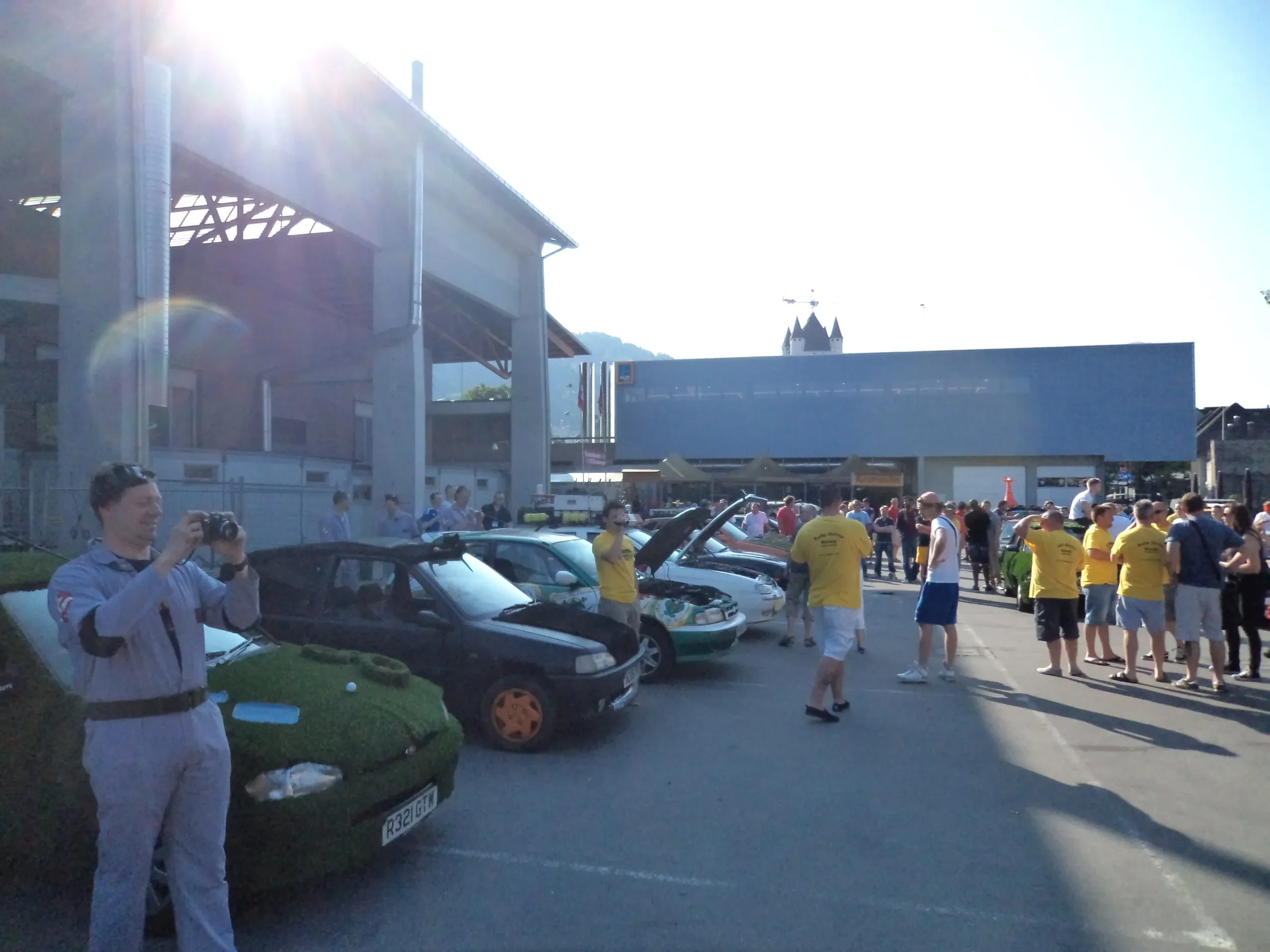 A crowd of people gathered outside a modern building with sports cars on display, some with hoods open, in a parking lot on a sunny day.
