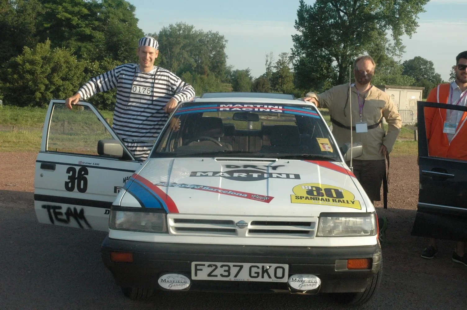 Three men standing around a white Nissan rally car with racing decals, one man in striped prison uniform, and two others wearing casual jackets, outdoors on a dirt road with trees in the background.