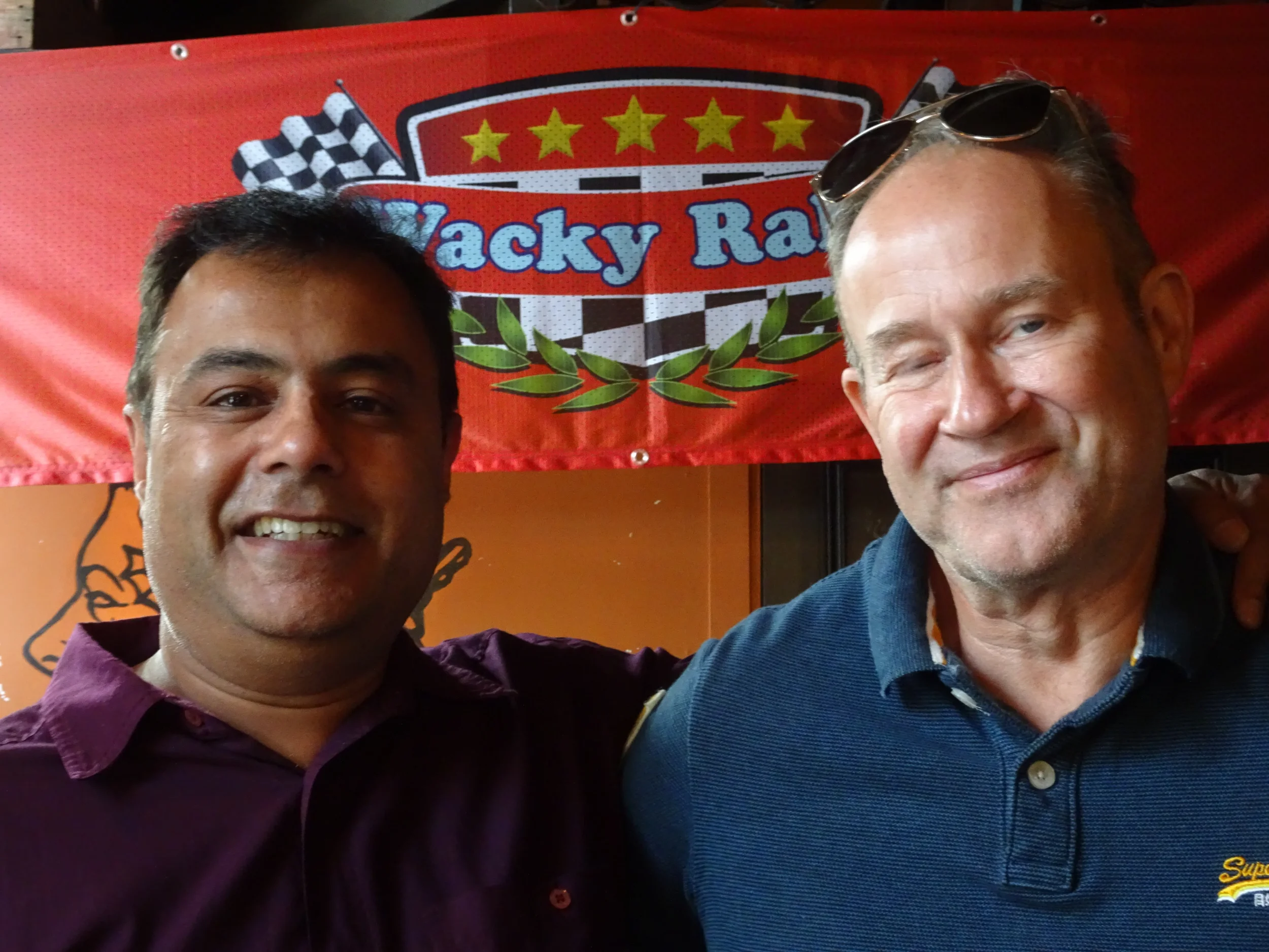 Two men smiling and posing for a photo in front of a red banner with a NASCAR-themed design and the words 'Macky R' visible.