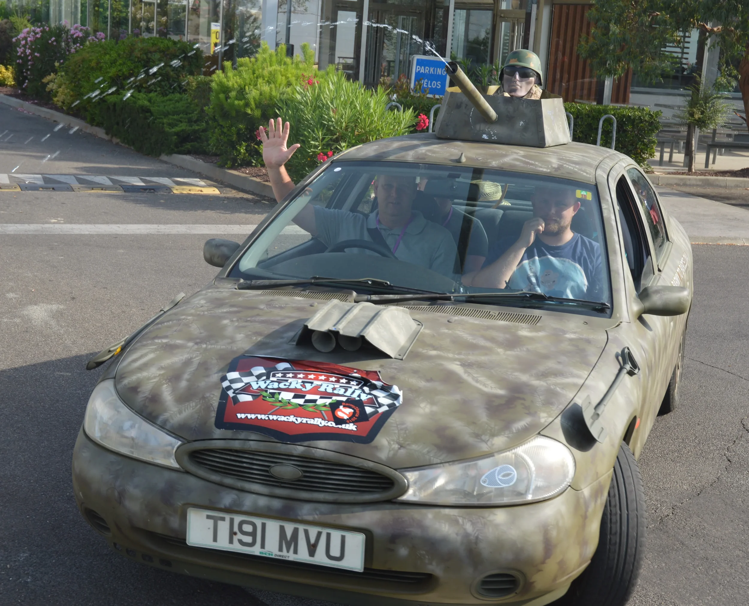 A small camouflage-colored car with a man waving and another man talking on the phone inside. The car has a logo on the hood and a humorous exhaust pipe. It is driven on a city street with plants and a sign in the background.