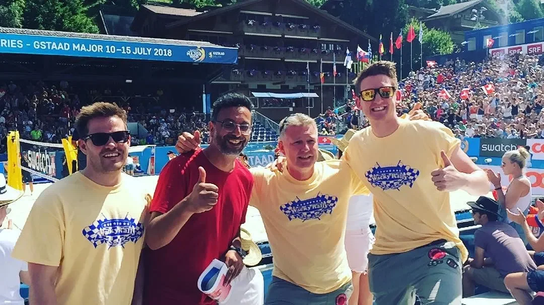 Four men at a sporting event, two wearing yellow T-shirts with a racing logo, one in a red T-shirt, and one in a white T-shirt; they are smiling and cheering, with a large crowd and flags in the background.