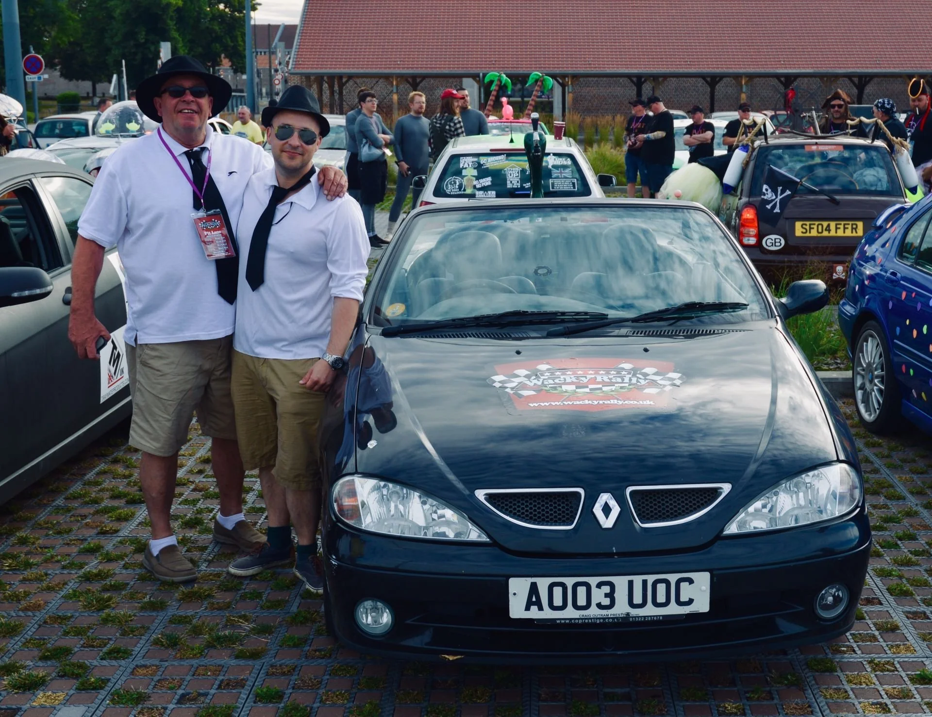 Two men dressed in white shirts, ties, and sunglasses standing together next to a black Renault car at a car event, with other decorated cars and people in the background.