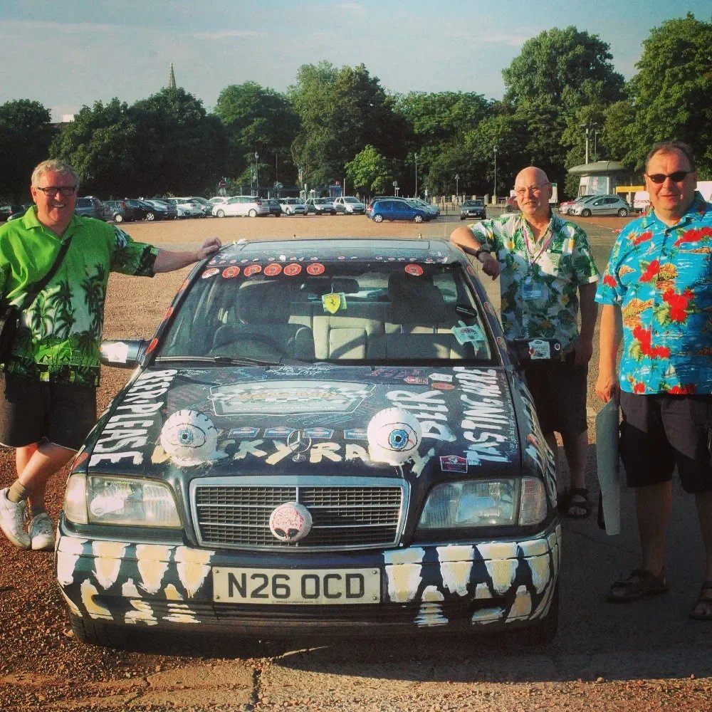 Three men in colorful tropical shirts standing next to a decorated car in a parking lot with trees and cars in the background.