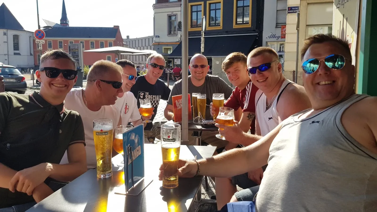 A group of nine men sitting at an outdoor table of a cafe, holding glasses of beer, smiling, and enjoying the sunny day.