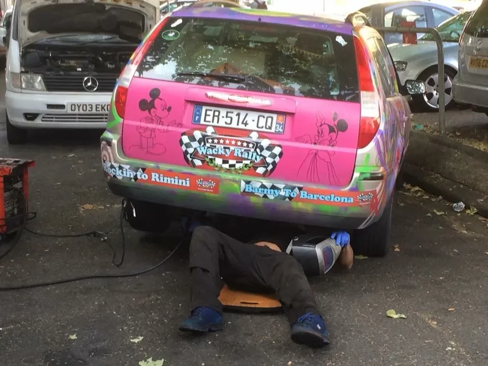 A person lying under a pink and purple car parked in a parking lot, performing car maintenance or repair. The car has decorations and stickers, including Mickey Mouse and Minnie Mouse, and promotional text for a rally from Rimini to Barcelona.