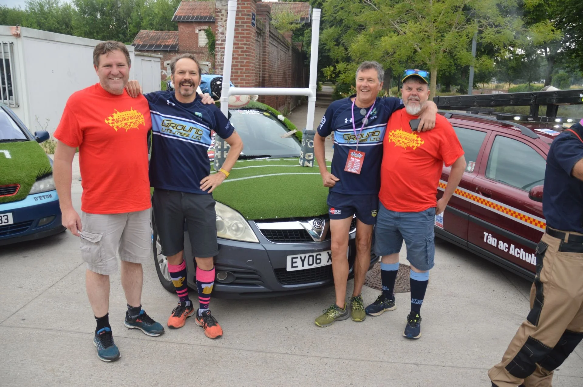 Five men standing together outdoors, smiling. Two are wearing red shirts, two are in blue shirts with 'Ground Utilities Ltd' logos, and one is in navy shorts. They are next to a car with a green grass-like hood, a vehicle with a red and yellow checke