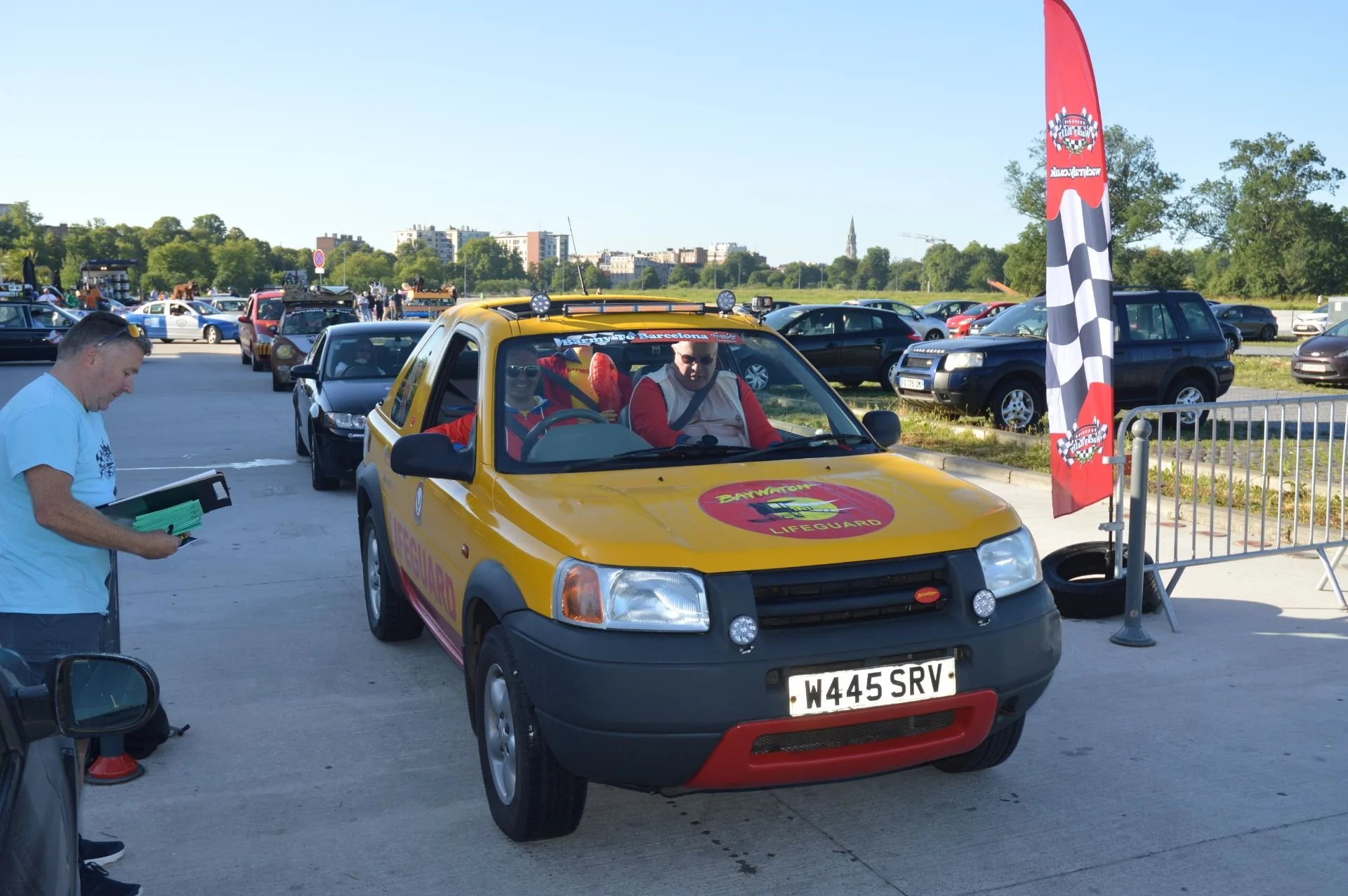 Yellow and black lifeguard vehicle at a race event with a man and woman inside, a man is standing outside reading a document, parked cars in the background, and a checkered flag on the side.