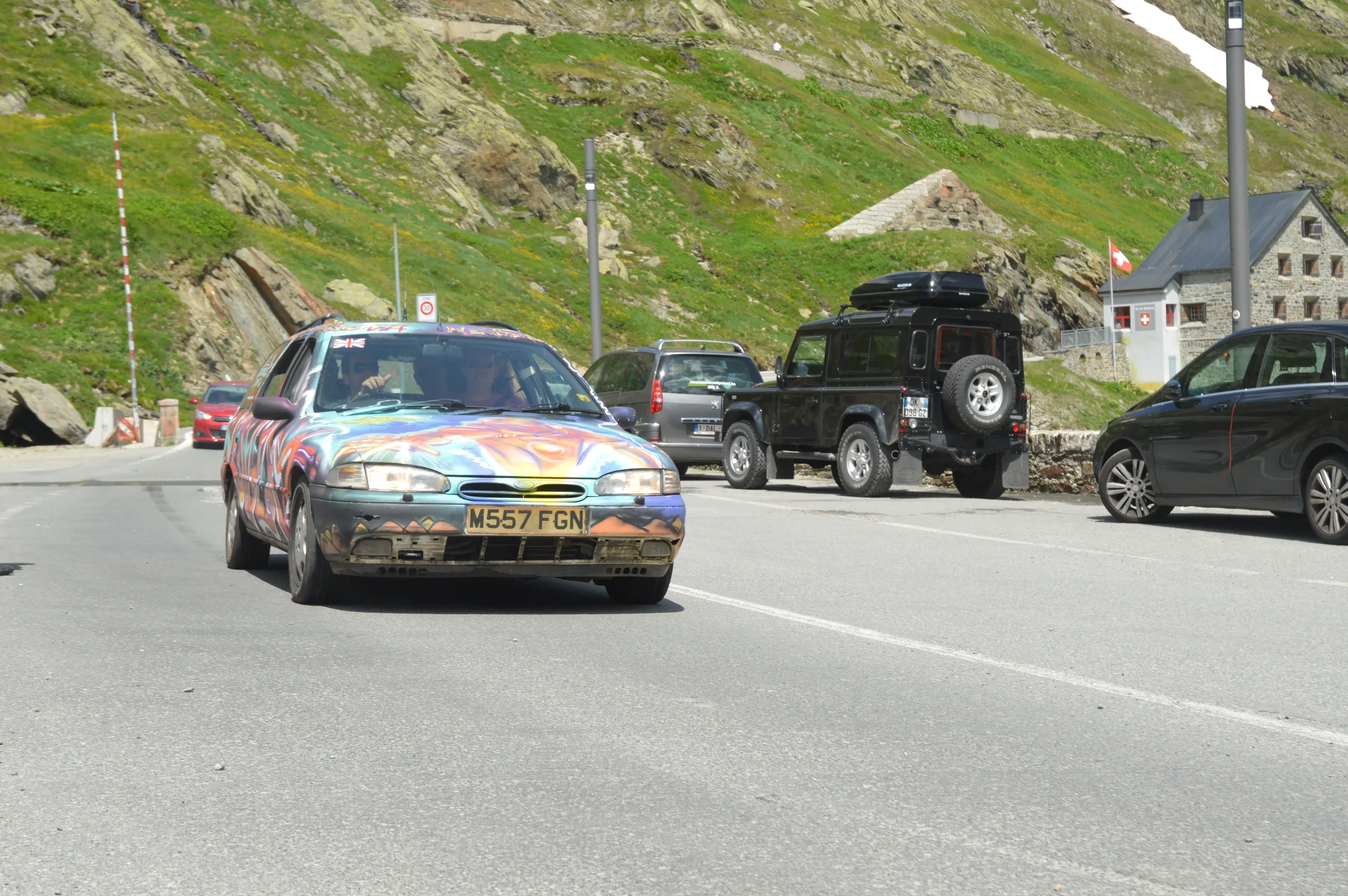 A parking area with several cars, including a colorful patterned car in the foreground, a black off-road vehicle in the middle, and a dark sedan to the right, with green rocky hills and a small stone building in the background.