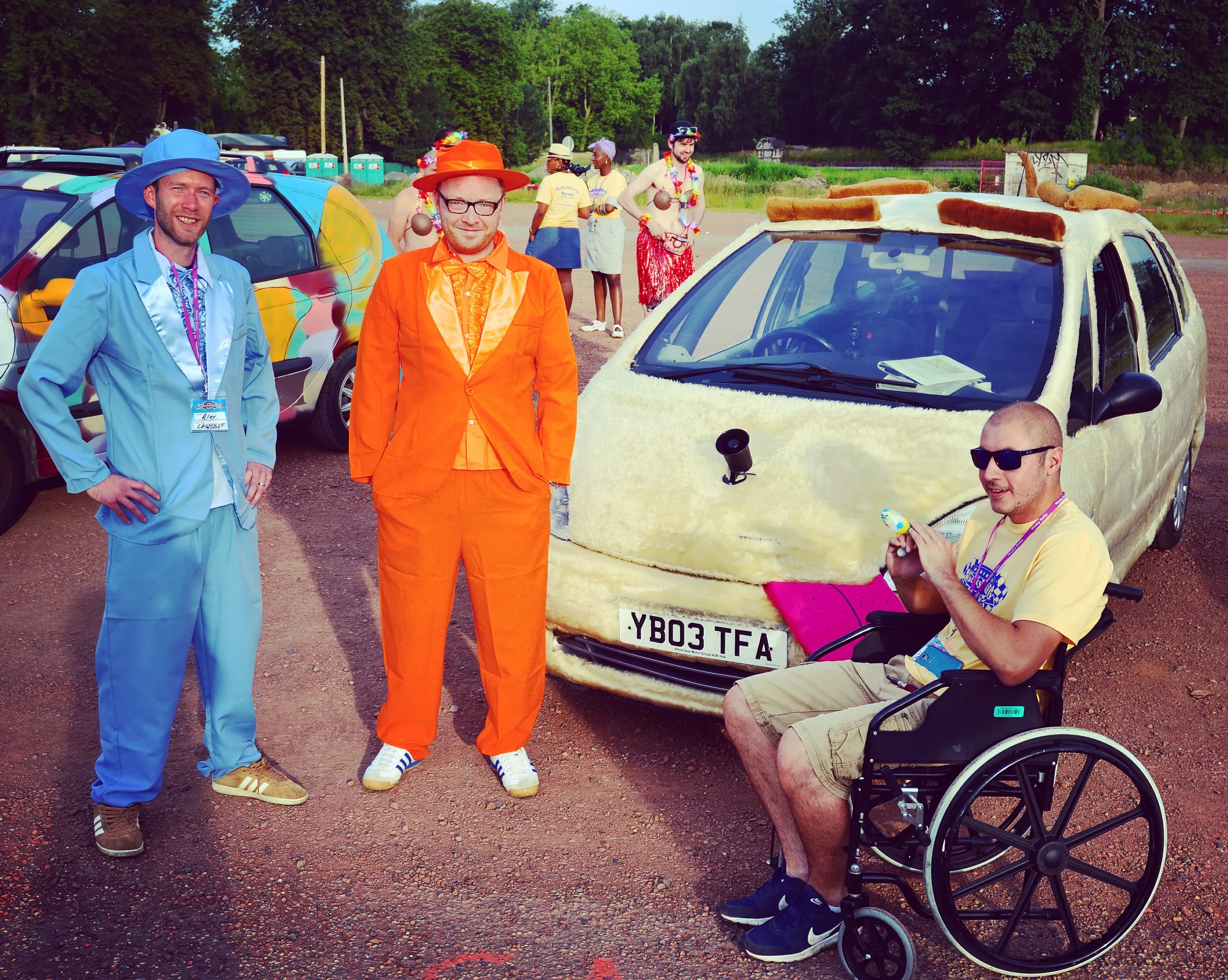 People dressed in colorful costumes standing next to a car covered in fluffy material resembling a sheep, with a man in a wheelchair in front of it, holding a small object.