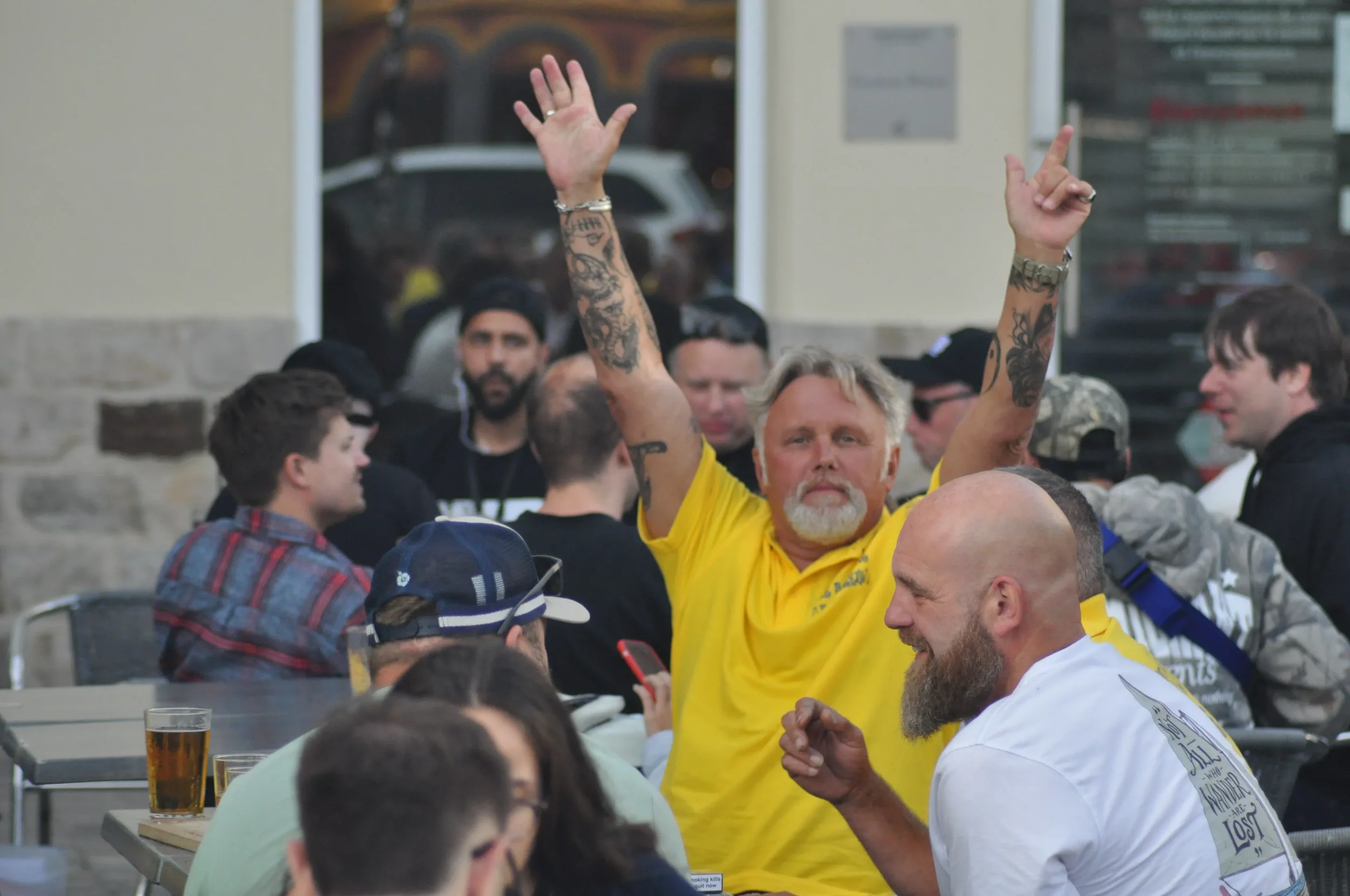 A group of people sitting at outdoor tables, with a man in a yellow shirt raising his arms in the air, and others chatting and smiling, some holding drinks.