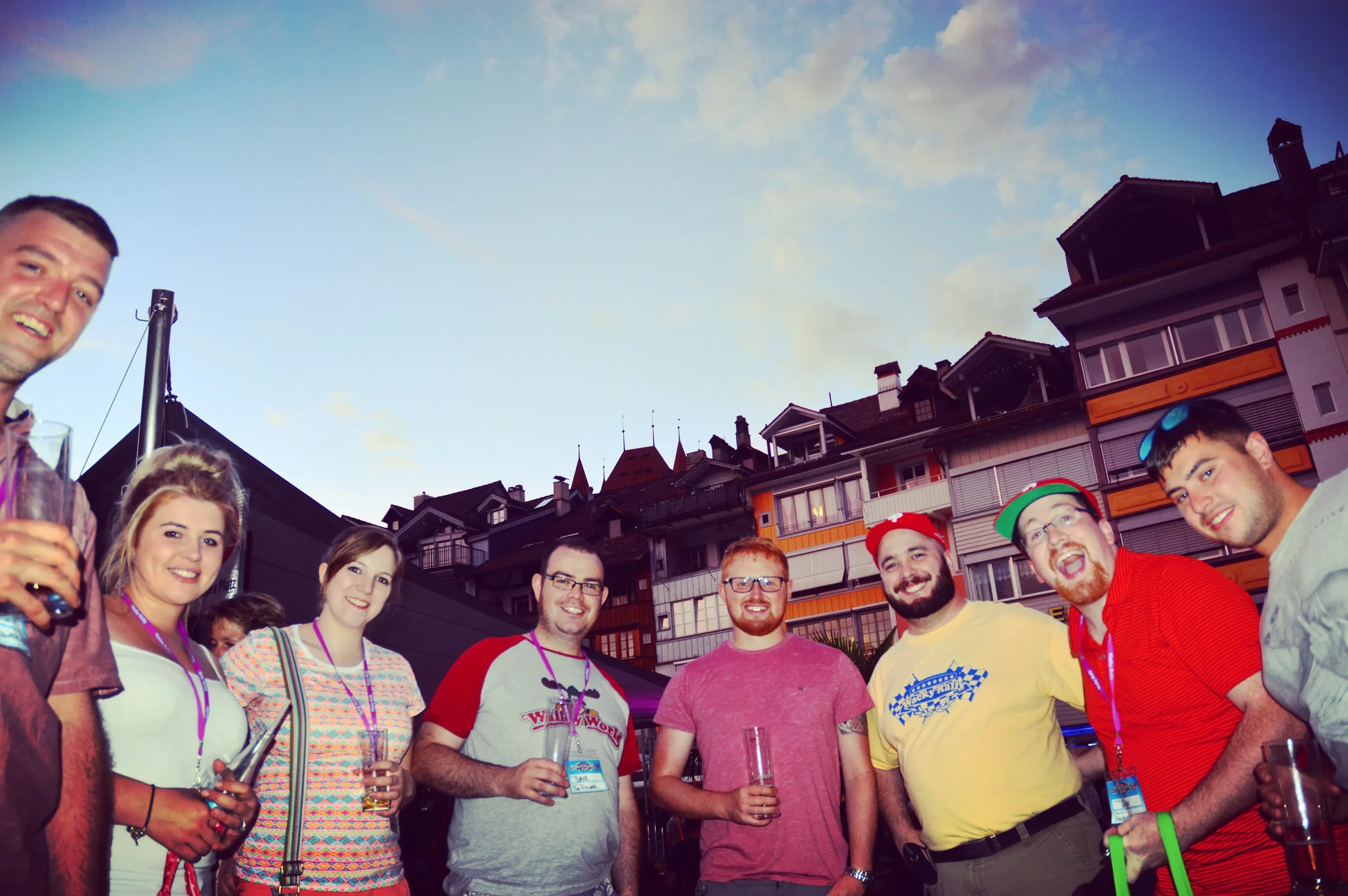 A group of eight young adults smiling and holding drinks at an outdoor event during the evening, with residential buildings in the background and a partly cloudy sky.