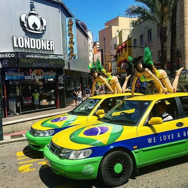 Two cars decorated in the colors and emblem of the Brazilian flag, with two women in samba costumes on top of each car, and a city street background with stores and pedestrians.
