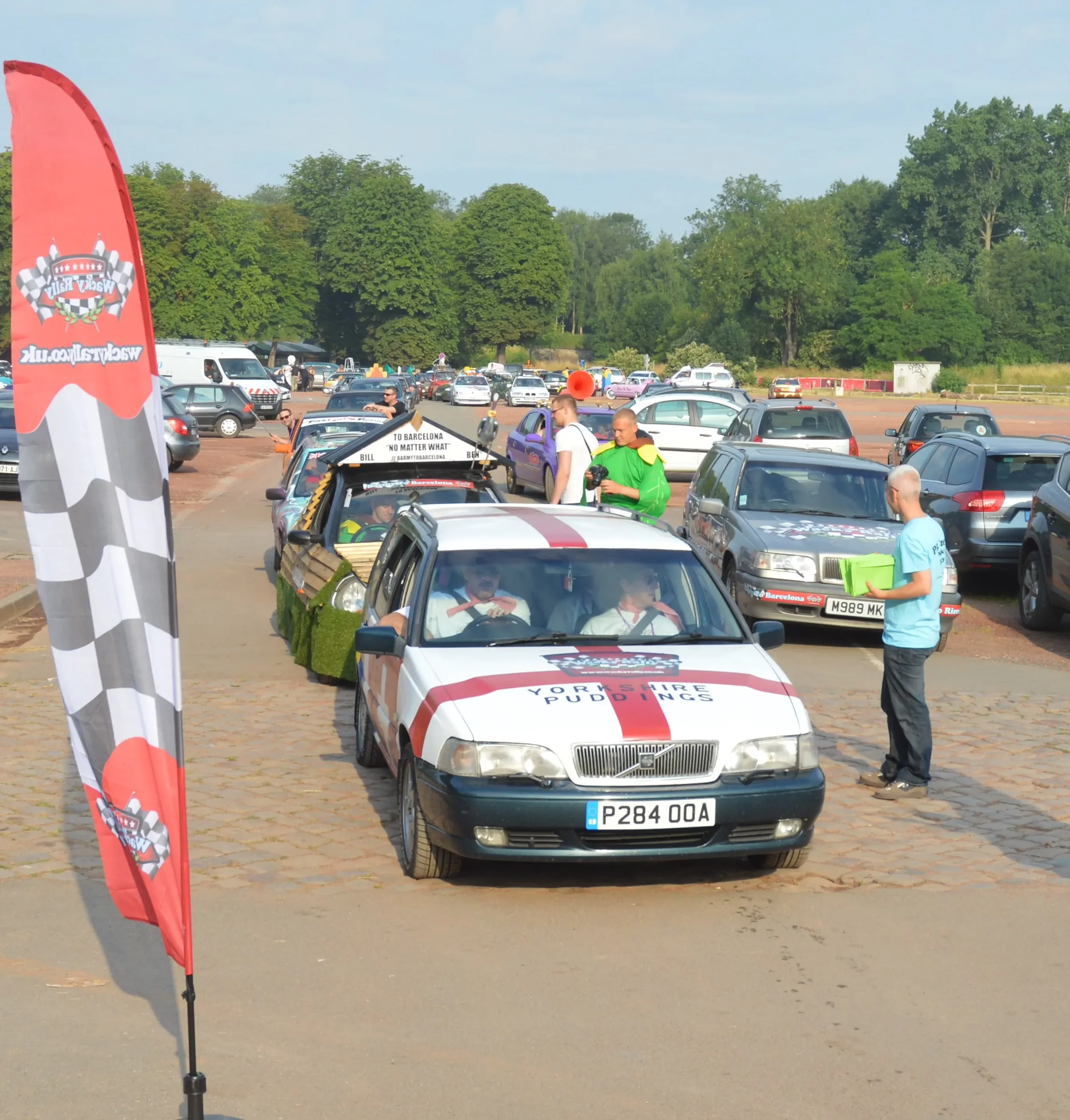 Line of decorated cars at a racing event with people gathered around, a flag with checkered pattern and racing logo, and a parking lot with green trees in the background.
