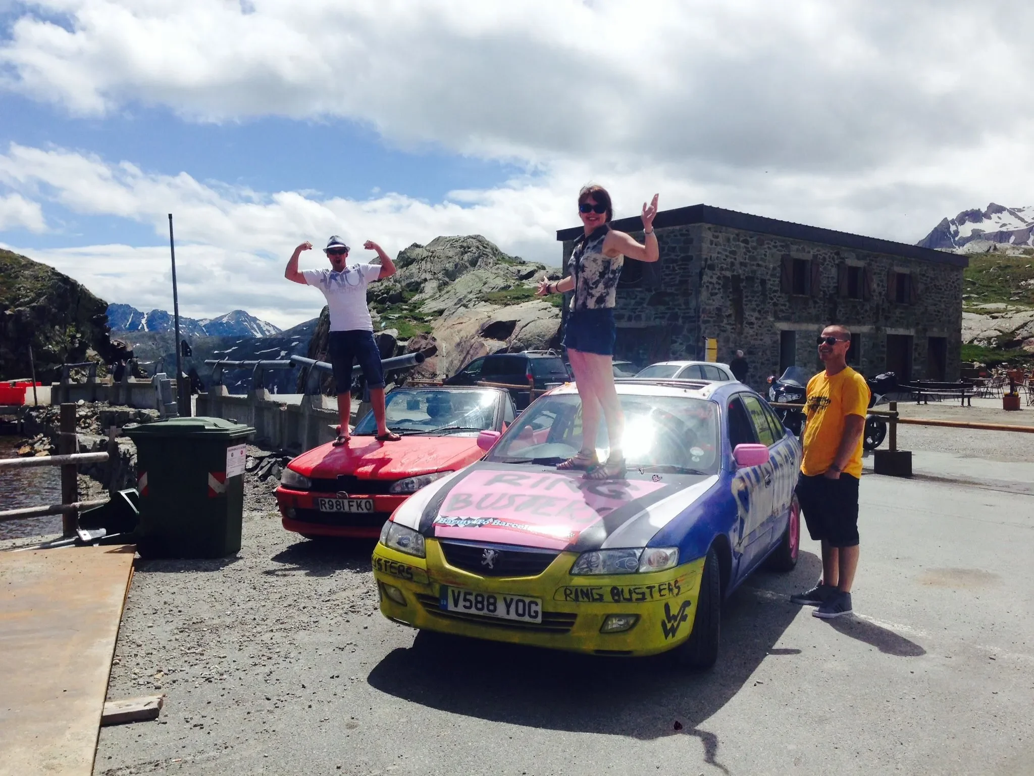 Three people are posing with cars in a mountainous outdoor setting with clouds and snow-capped peaks. One woman stands on a colorful car with "Ring Busters" written on it, another woman stands on a red car flexing her muscles, and a man stands beside