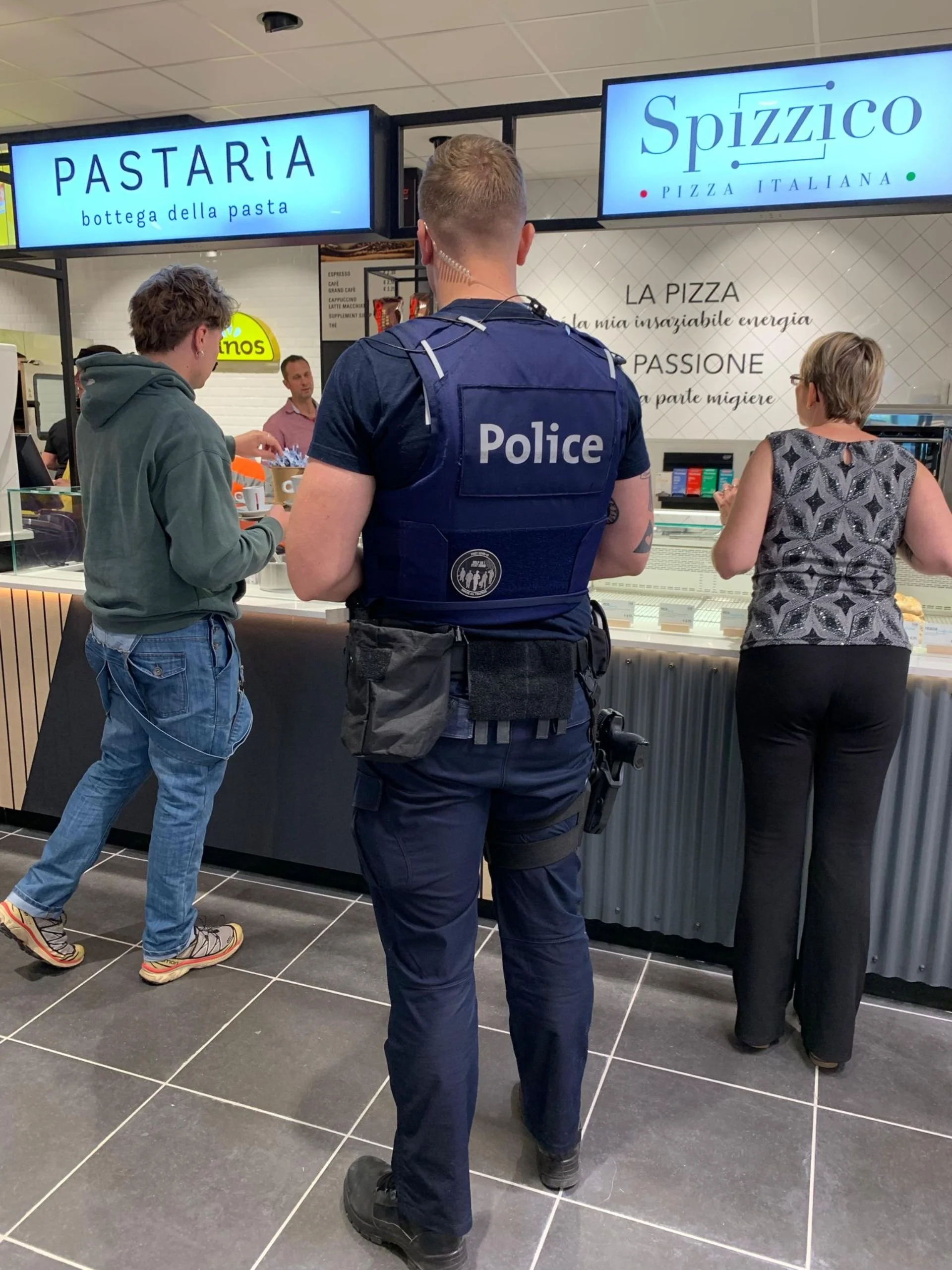 A police officer standing at a counter in a pizza shop with customers, including a young man and an older woman, ordering food.