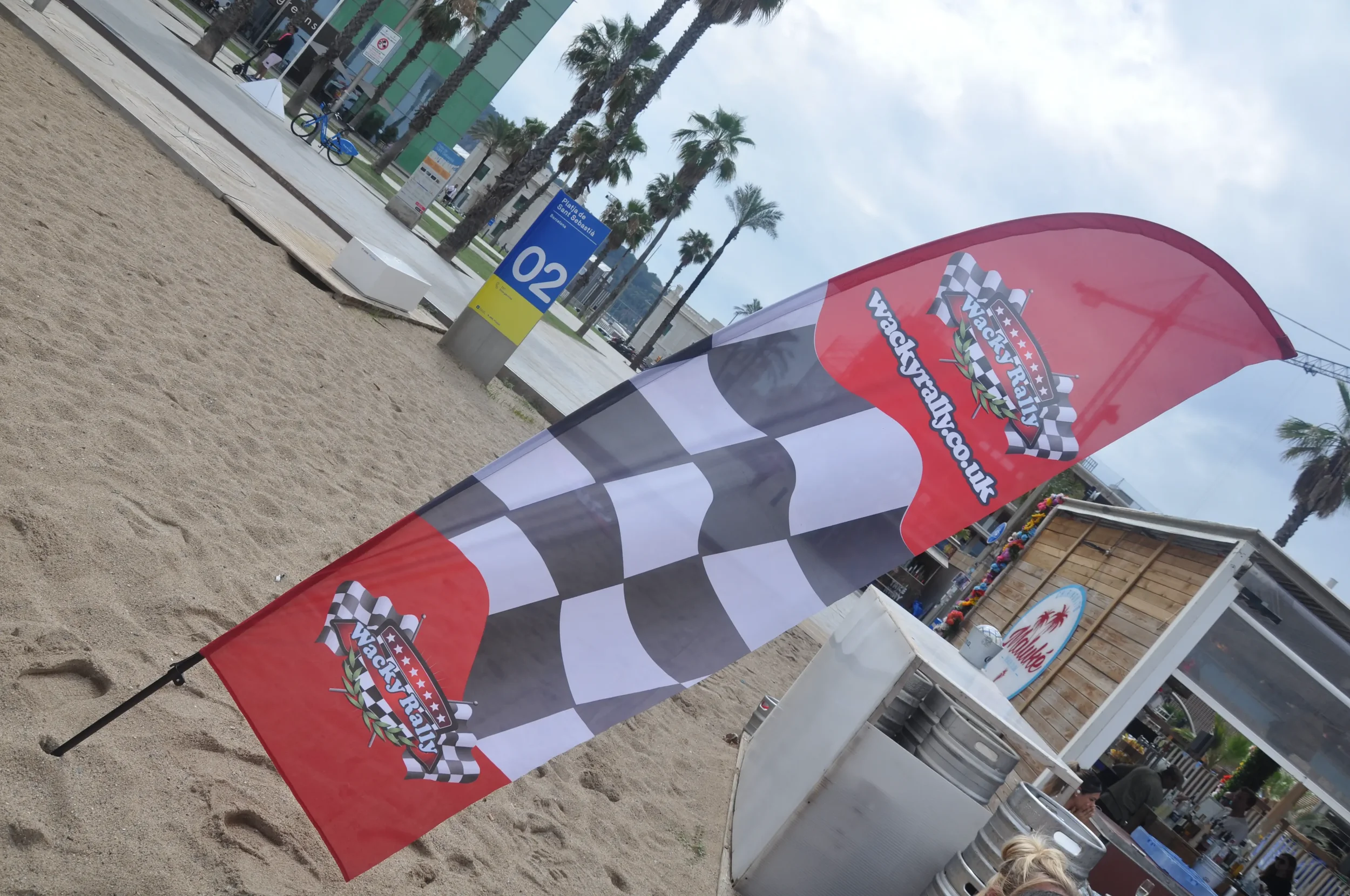 A red and black flag with chequered pattern and 'Wacky Races' logo standing on sandy ground at an outdoor event with palm trees and a blue sky in the background.