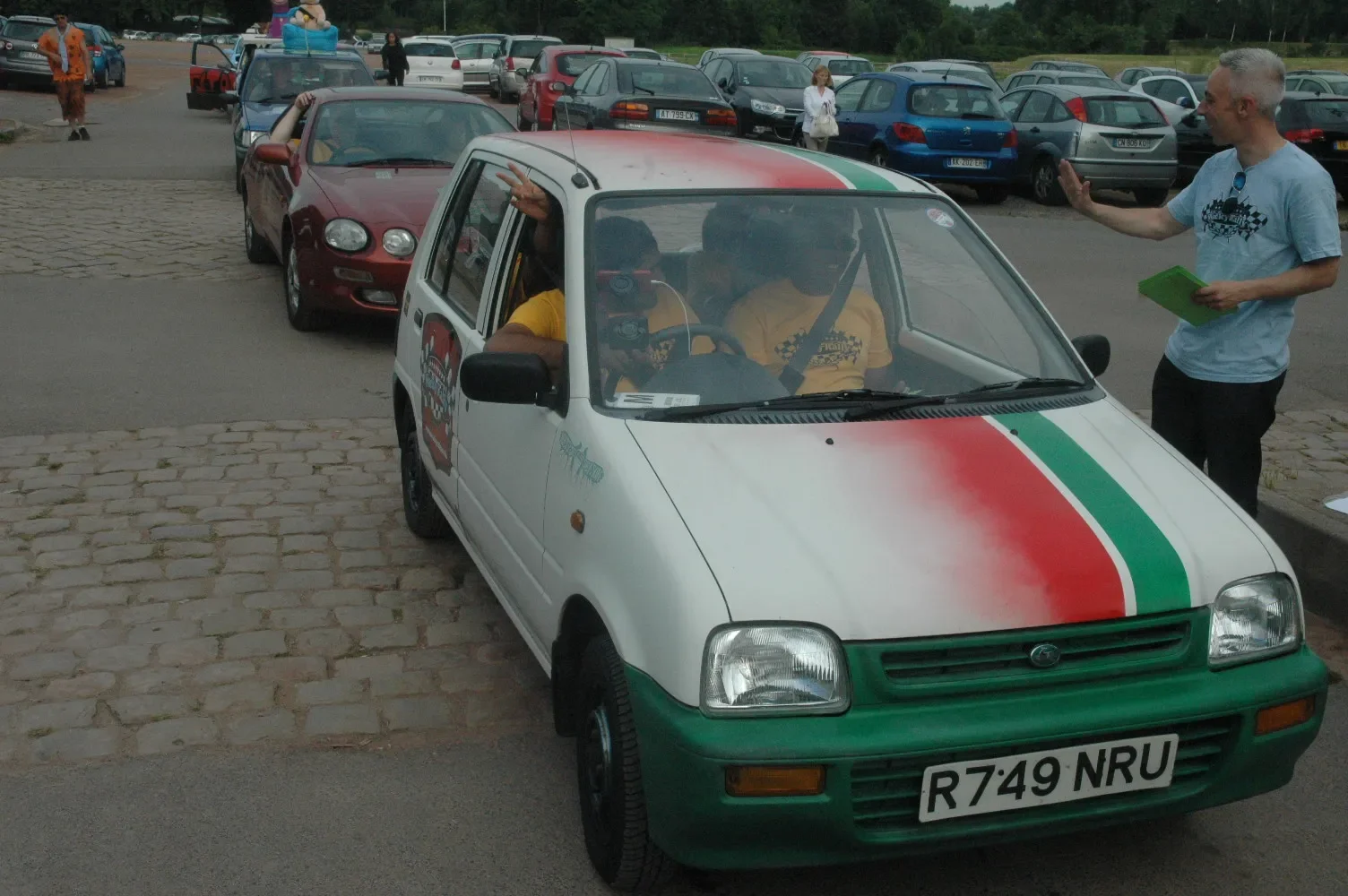 A small white car with Italian flag colors on the hood, parked in a lot with other cars, with two people inside and a man standing outside talking to them, holding a green notebook.