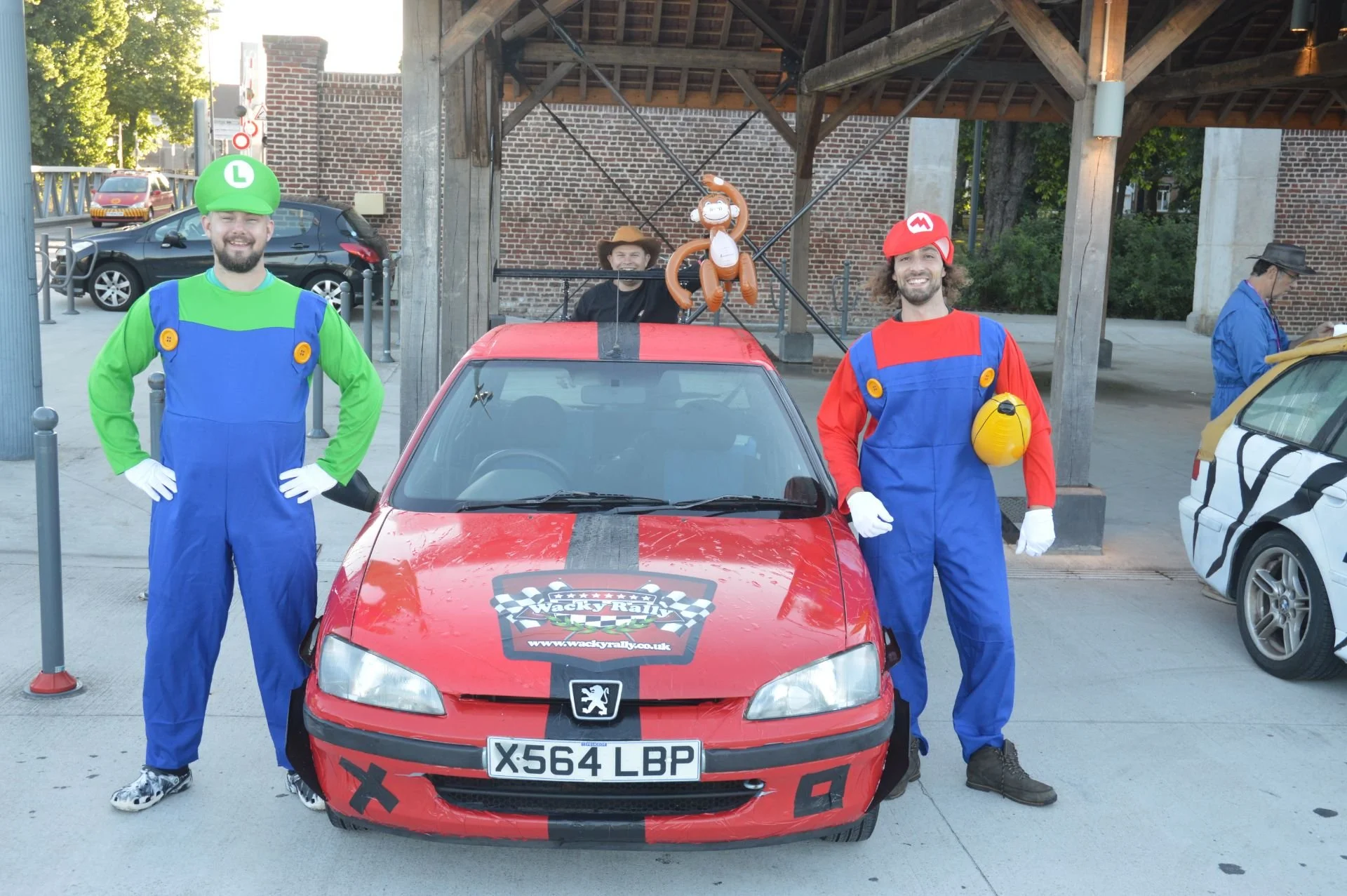 Two men dressed as Mario and Luigi from Mario Kart standing next to a red car with a Pokémon balloon on top, under a wooden structure in an outdoor setting.