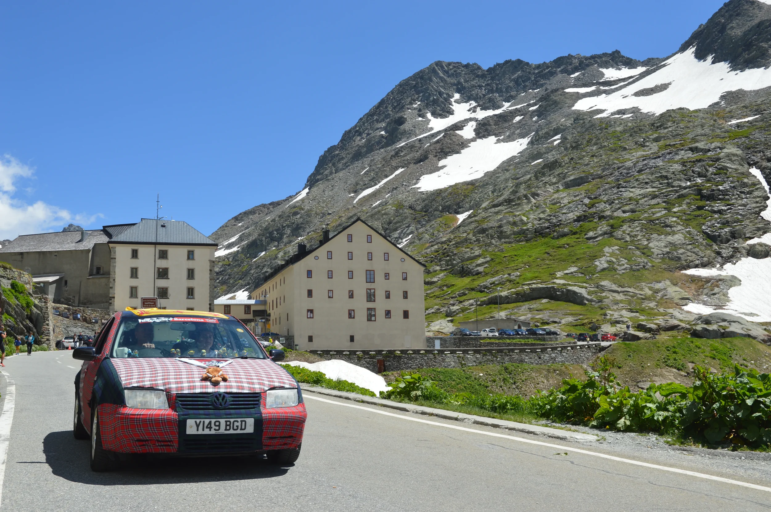 A car with red plaid wrapping and stuffed animal on hood parked on mountain road with large yellow building and snow-capped mountain in background.
