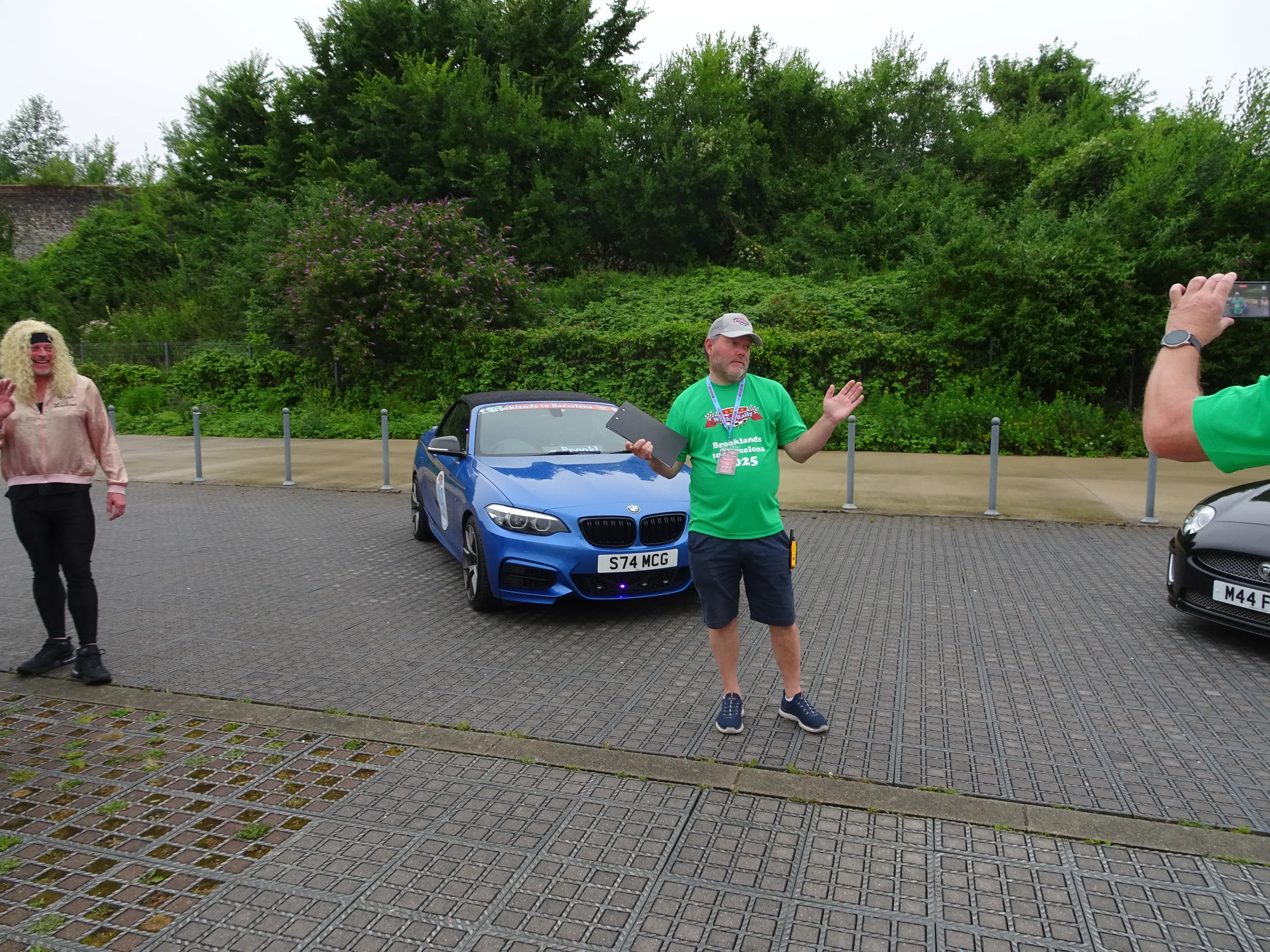 Three people standing next to cars in a parking lot, with one person in a green shirt holding a clipboard and another person taking a photo, in front of a green, leafy background.