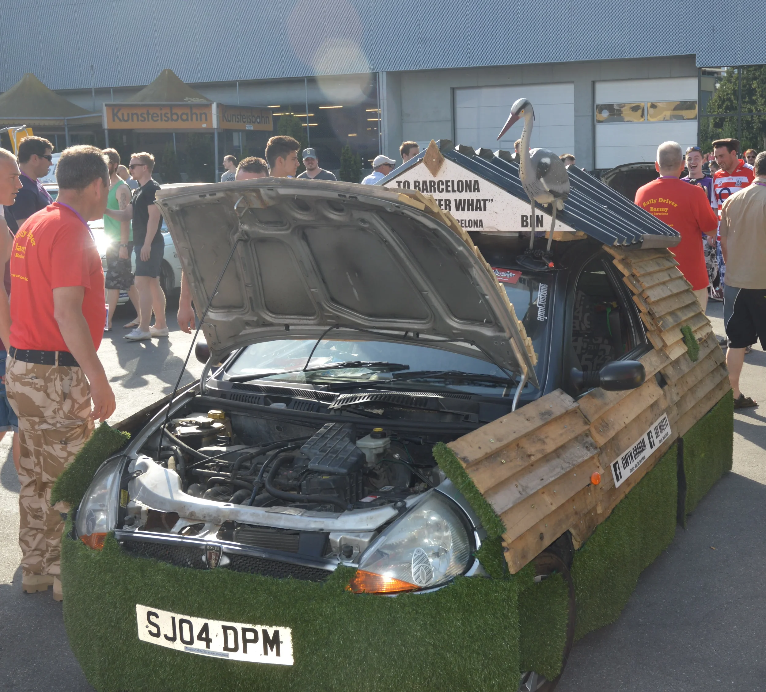 Car decorated with artificial grass, wooden slats, and a swan figure on top, displayed at a public event with people observing.