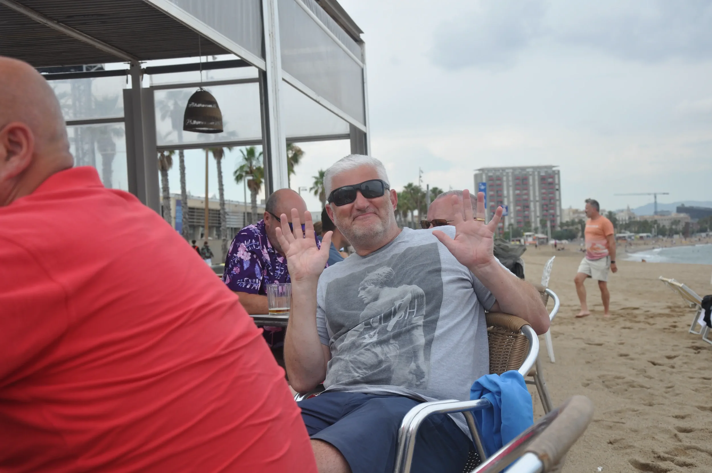 A man with gray hair and sunglasses sitting on a chair at a beachside restaurant, smiling and raising both hands, with beach and city buildings in the background.