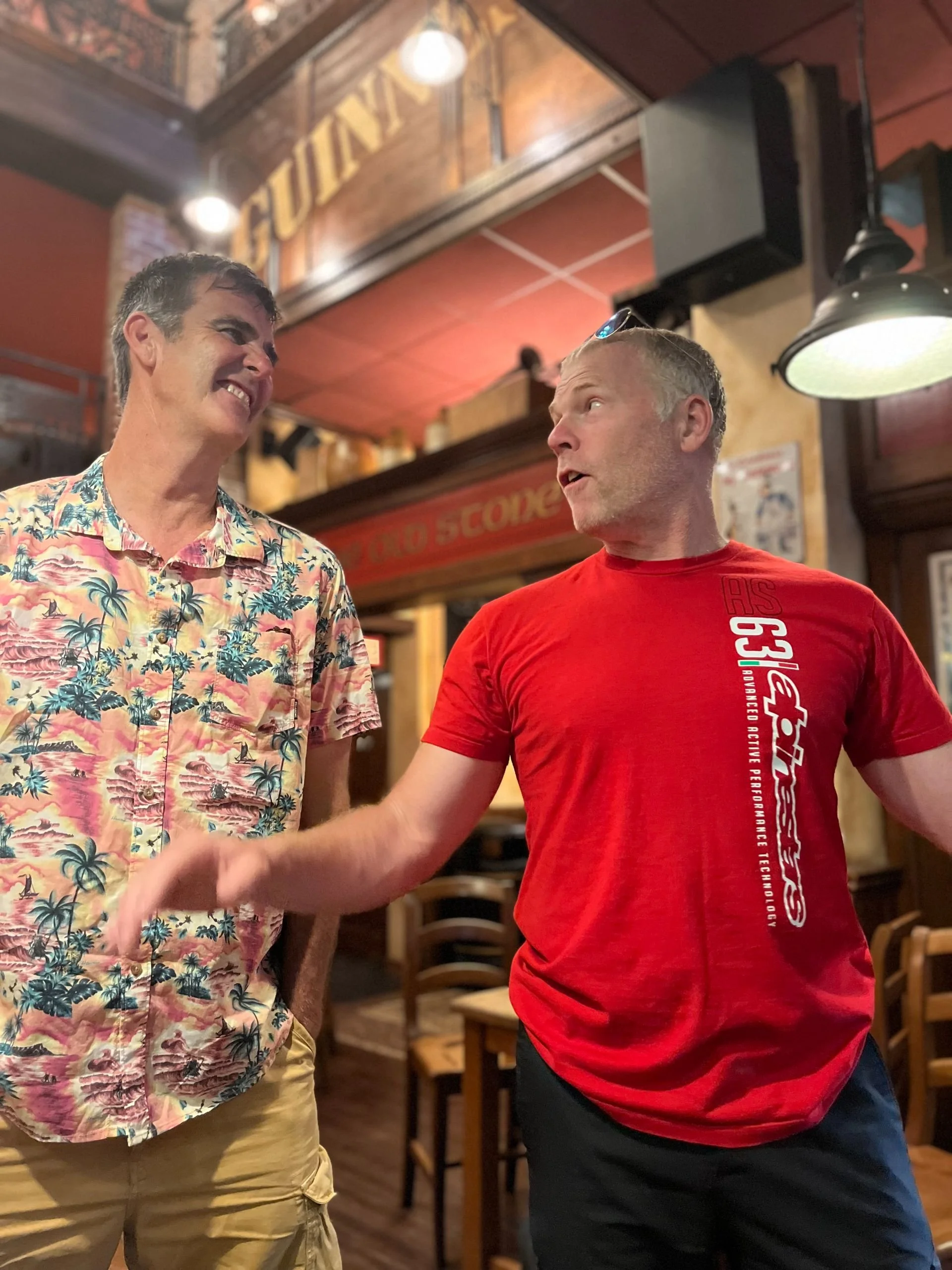 Two men are having a lively conversation in a bar or restaurant. One is wearing a colorful Hawaiian shirt, and the other is in a red T-shirt. They appear animated and expressive, with the man in the red shirt gesturing as he talks.