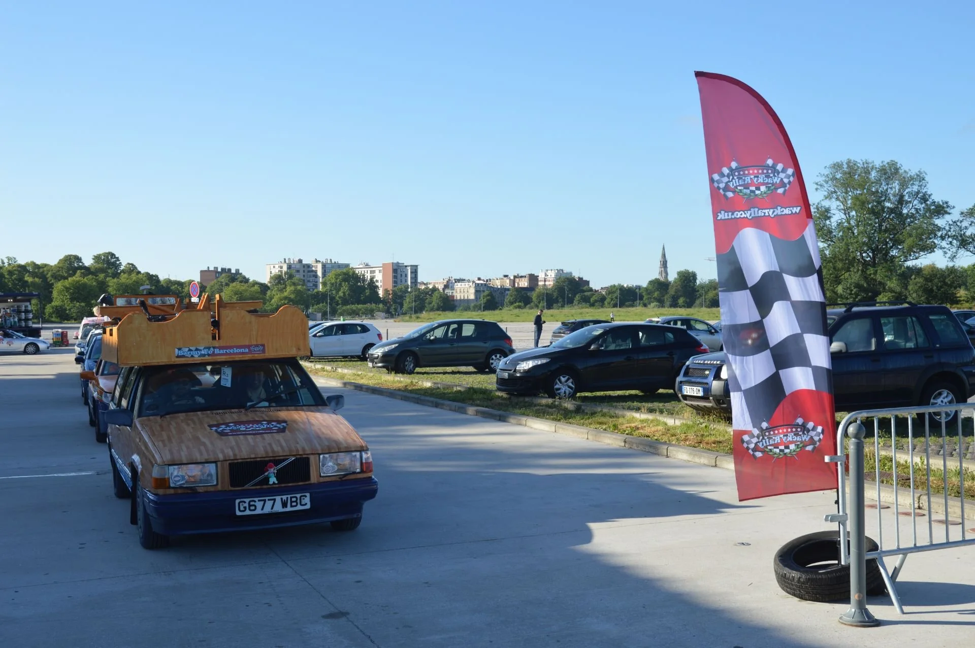A parking lot with several cars parked and a vintage car with a wooden exterior and a model of a decorative Christmas tree on the hood. There is a red flag with a racing flag pattern and the text "Vintage Car Club" on a stand. In the background, ther