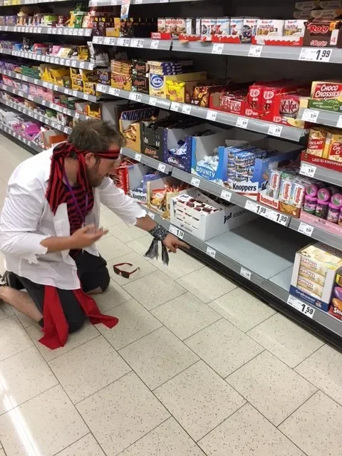 Person dressed as a pirate kneeling on supermarket floor pointing at snacks on a lower shelf.