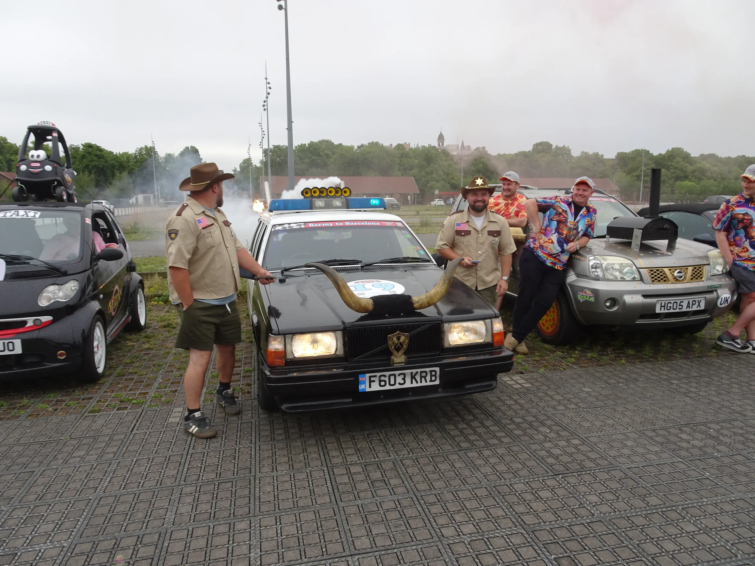 A group of people dressed in costumes, standing next to decorated cars, including a black car with a large horn on its hood and a police toy car with smoke and sparks in the background, at an outdoor event on a cloudy day.