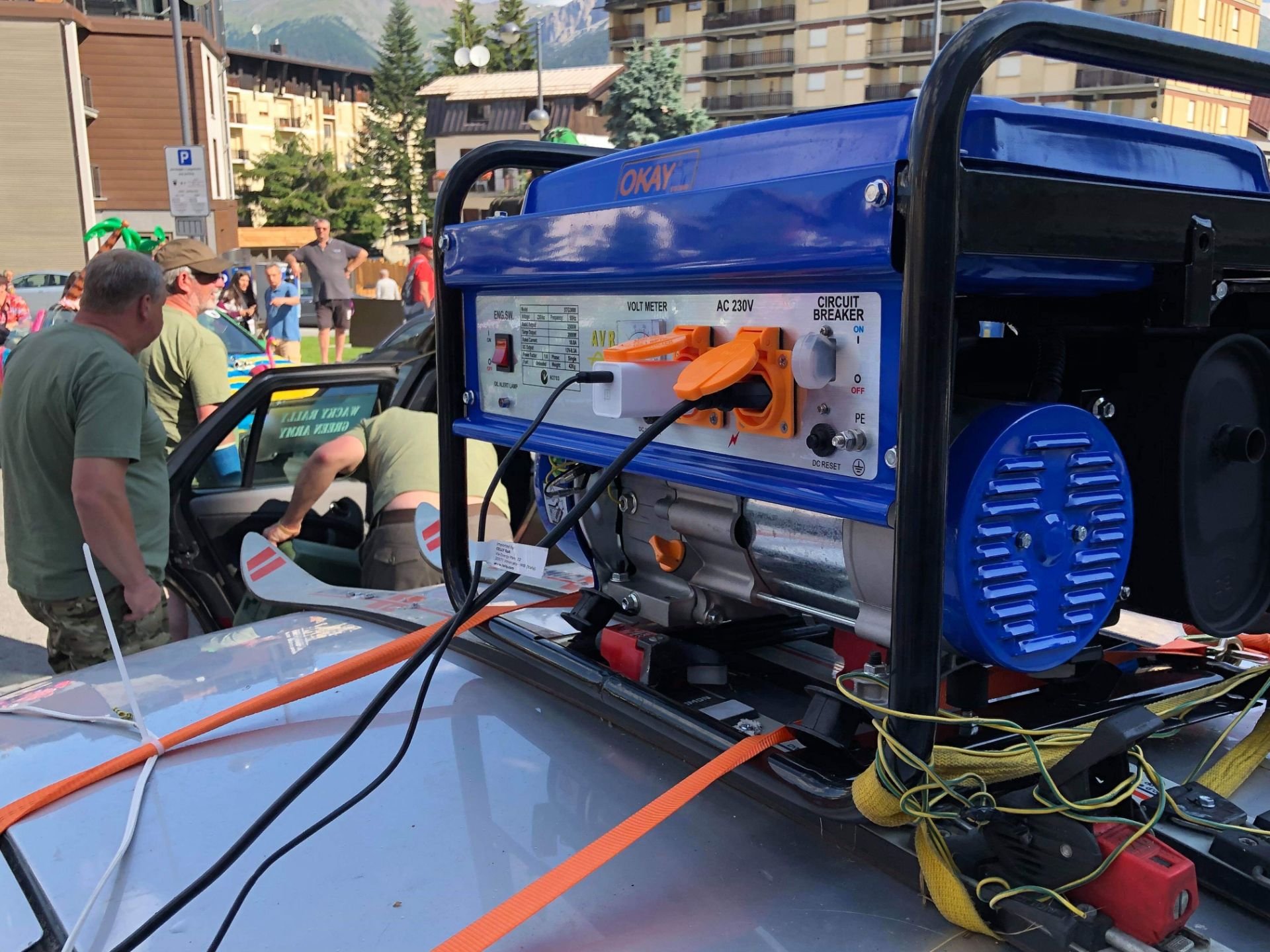 A close-up of a portable power generator placed on the roof of a vehicle at an outdoor event, with people and buildings in the background.