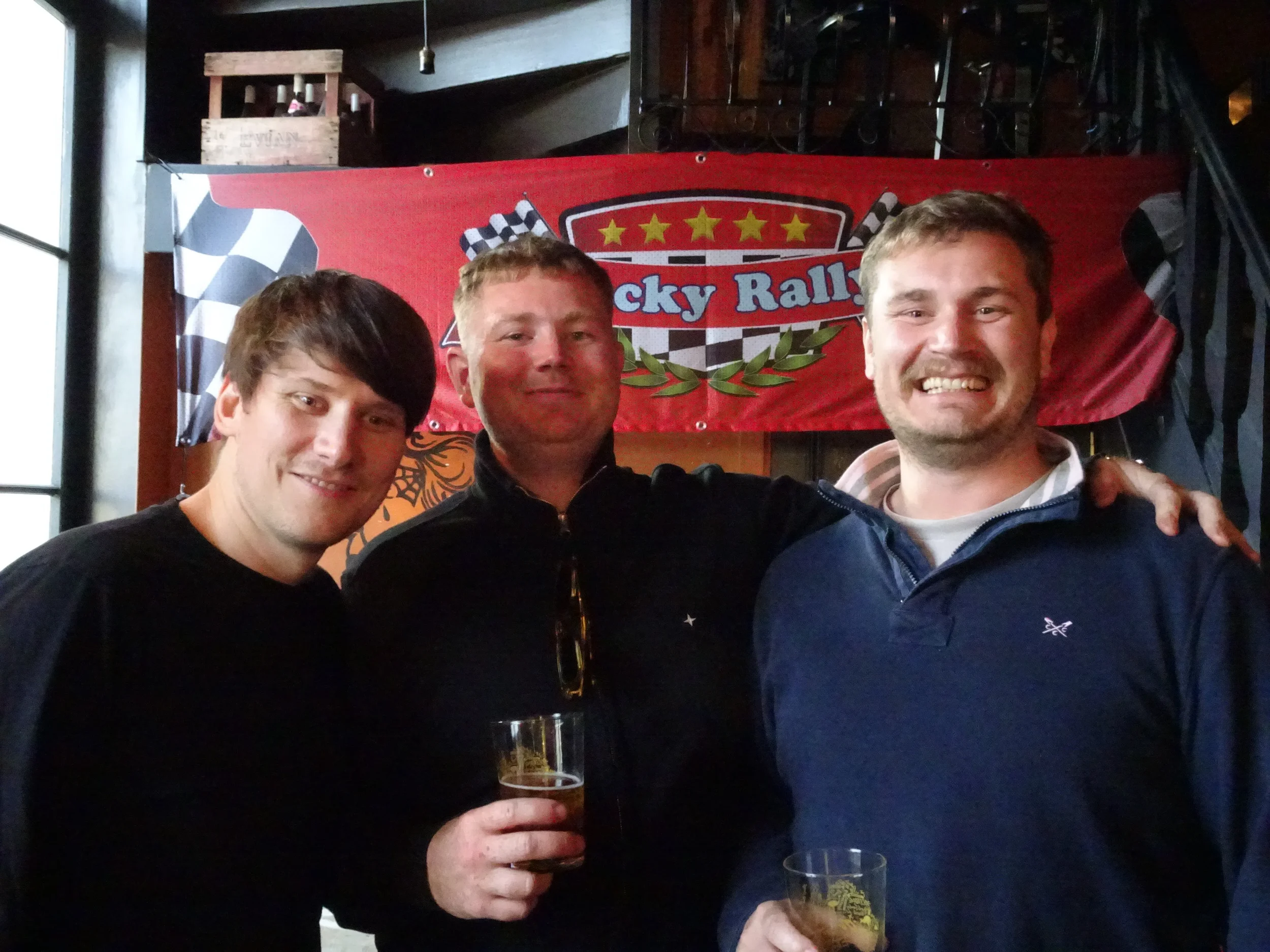 Three men smiling and holding drinks at a NASCAR-themed event, standing in front of a red banner with checkered flags and the words "Rocky Rally".