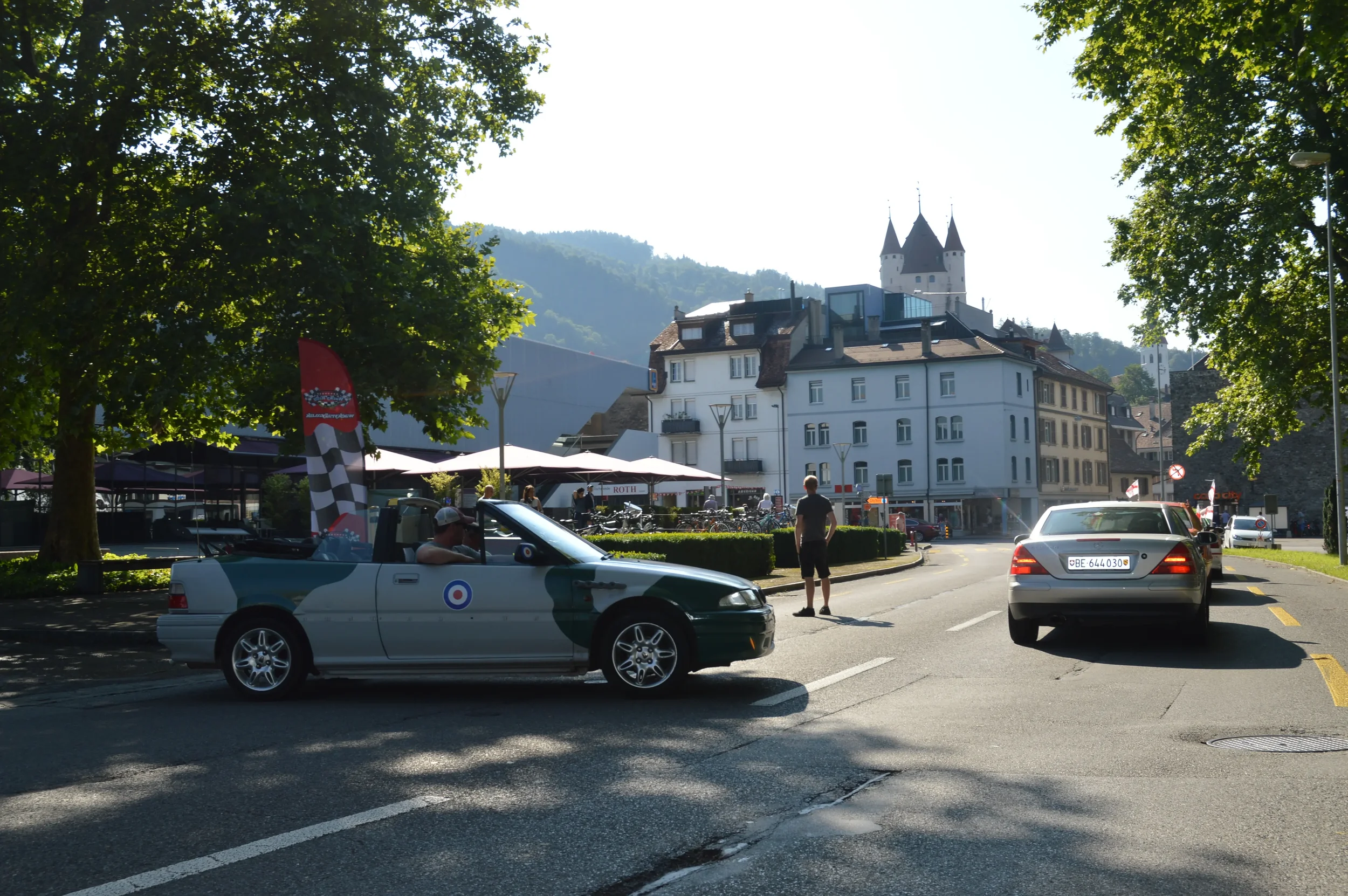 A street scene with a vintage green convertible car, a man standing on the road, another person sitting in the car, and multiple parked cars. In the background, there are white buildings, a hill with a castle, and trees on the sides.