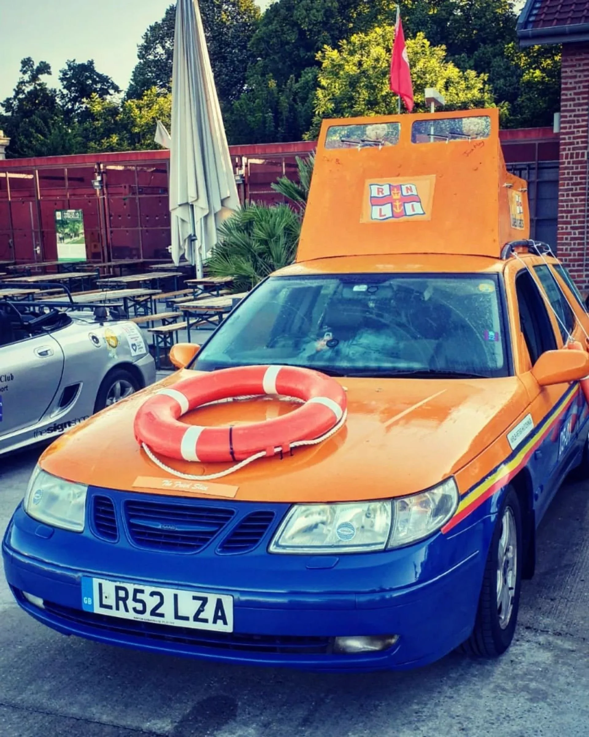 An orange and blue vehicle with a lifebuoy on its hood and a boat-shaped structure on top, decorated with various maritime symbols, parked in an outdoor area with tables, umbrellas, and trees in the background.