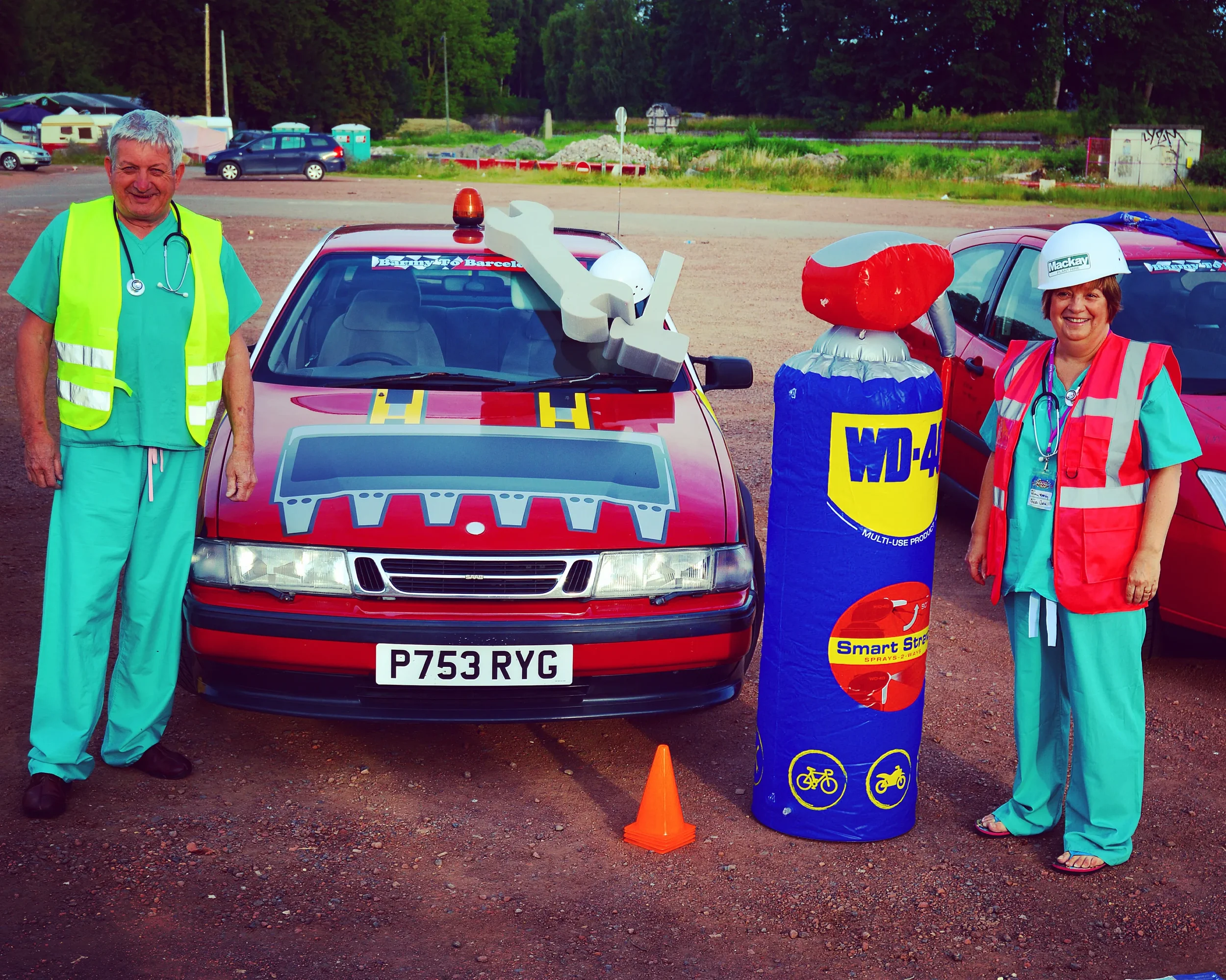 Two healthcare workers in scrubs and safety vests standing next to a red rescue or emergency vehicle with a large WD-40 can and a safety cone in front. The vehicle has a decal of a mechanical wrench on the hood, and the scene is set in a parking lot 