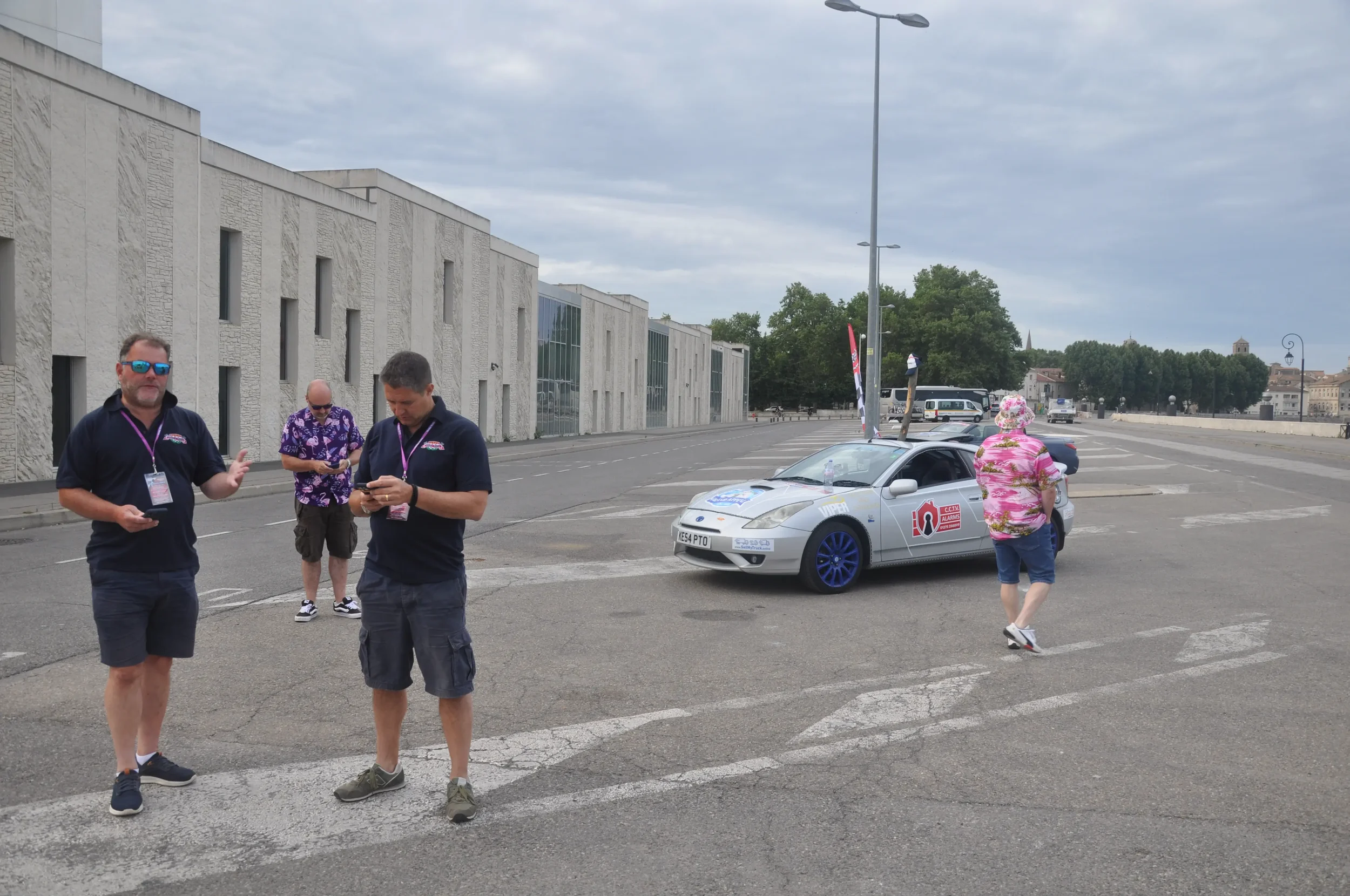 Four people outside near a silver sports car with travel or event logos in a paved open area, overcast sky, modern white building, trees, and parked buses in the background.
