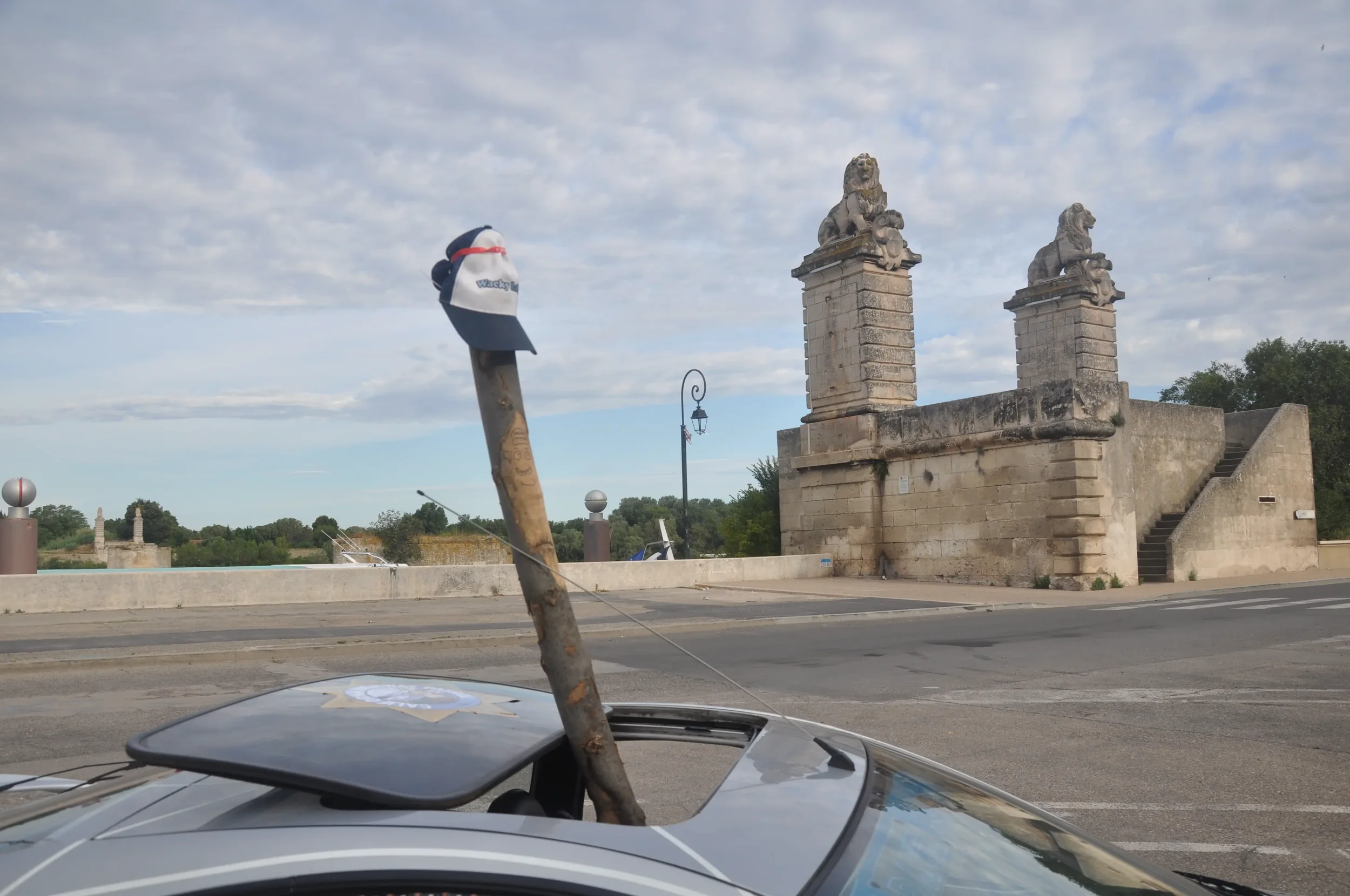 View of a historic stone bridge with two carved lion statues atop brick pillars, a parking lot in foreground, and a hat hanging on a stick attached to a car.