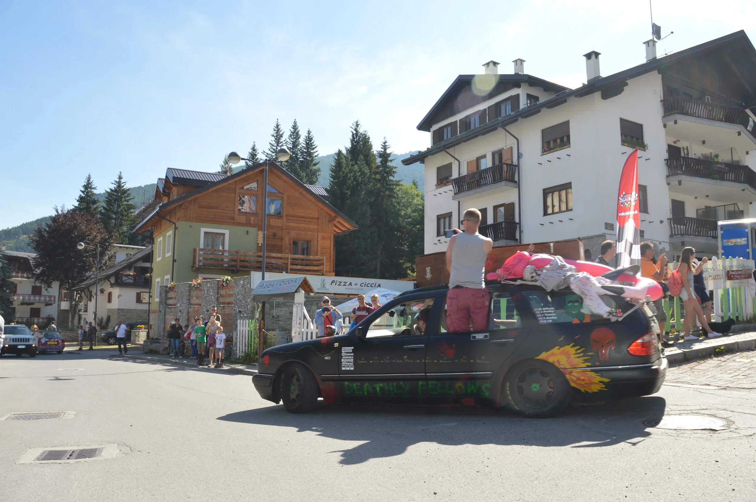 A black car with flames and red skull decals, labeled 'Deathly Fellows,' parked on a street in a mountain village, with a person standing on top, and groups of people gathered around.