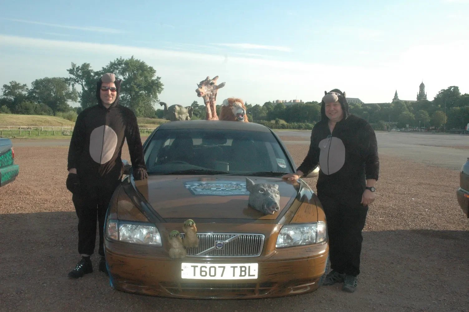 Two women dressed in animal costumes with hats resembling animals, posing outdoors next to a brown Volvo car decorated with animal plush toys and props, including a wolf's head on the hood and giraffe and lion puppets, with three animated animal mask