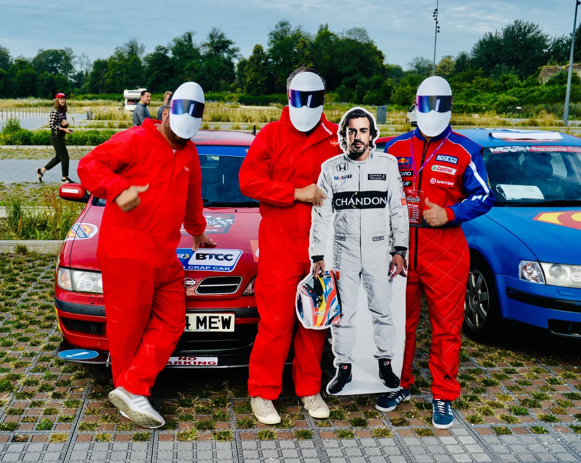 Three people in racing suits wearing helmets standing in front of cars, holding a life-sized cardboard cutout of a man in a racing suit, at a racing event.