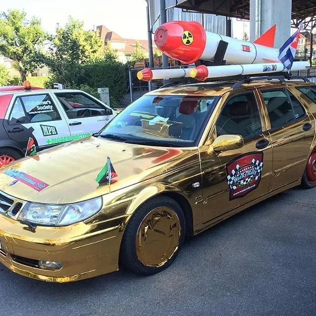A gold-colored car decorated to look like a racing car with a small flag on the front grille, a checkered flag emblem on the door, and a large missile model on the roof.