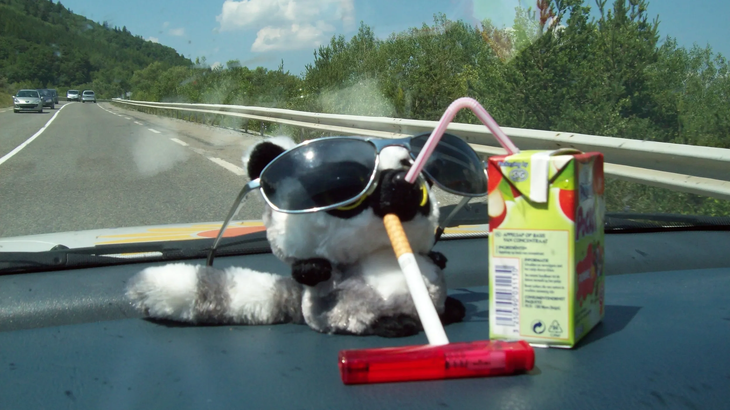 A stuffed panda bear toy wearing sunglasses, smoking a cigarette, with a juice box and straw on a car dashboard during a road trip.