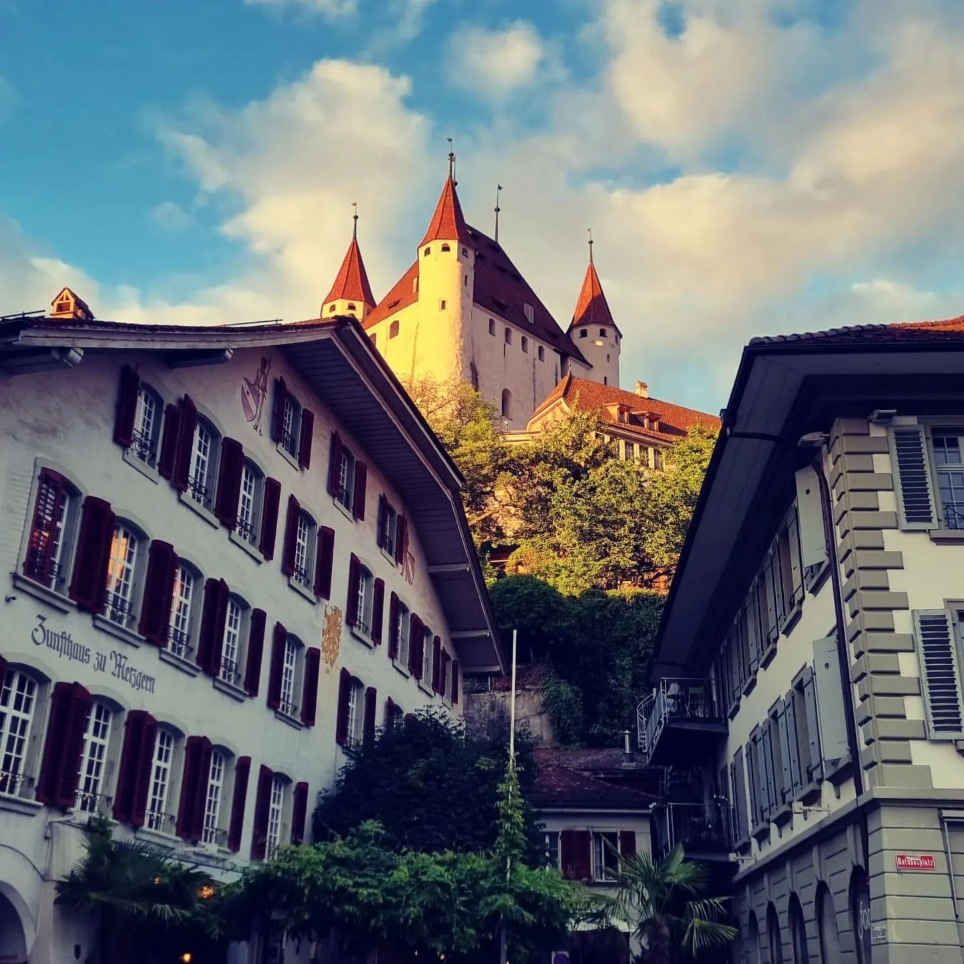 A castle with three pointed towers on a hill, surrounded by buildings with European architecture and tree-strewn streets.