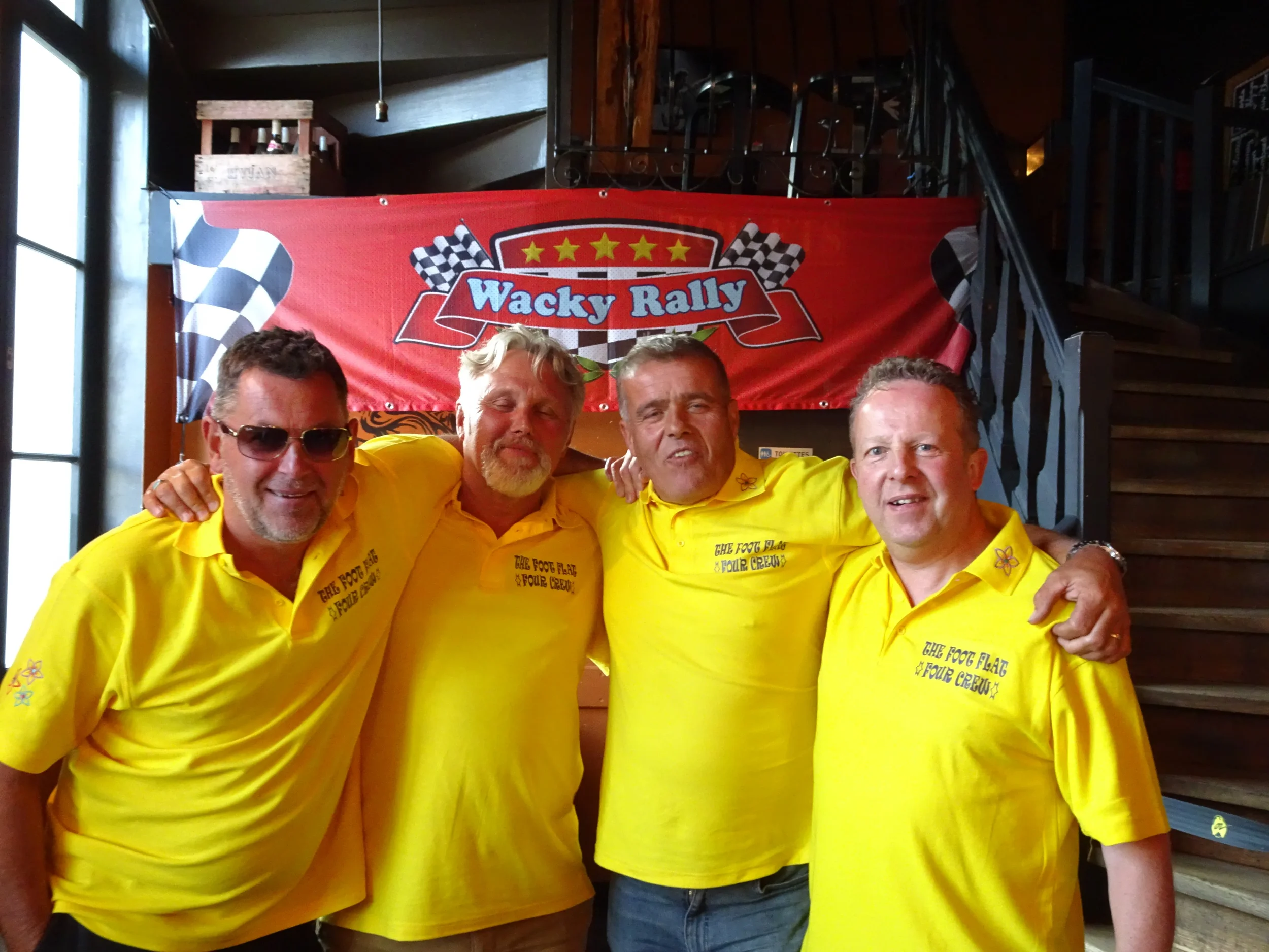 Four men in yellow shirts standing together at a rally event with a red banner behind them that reads "Wacky Rally." The men are smiling and have their arms around each other.