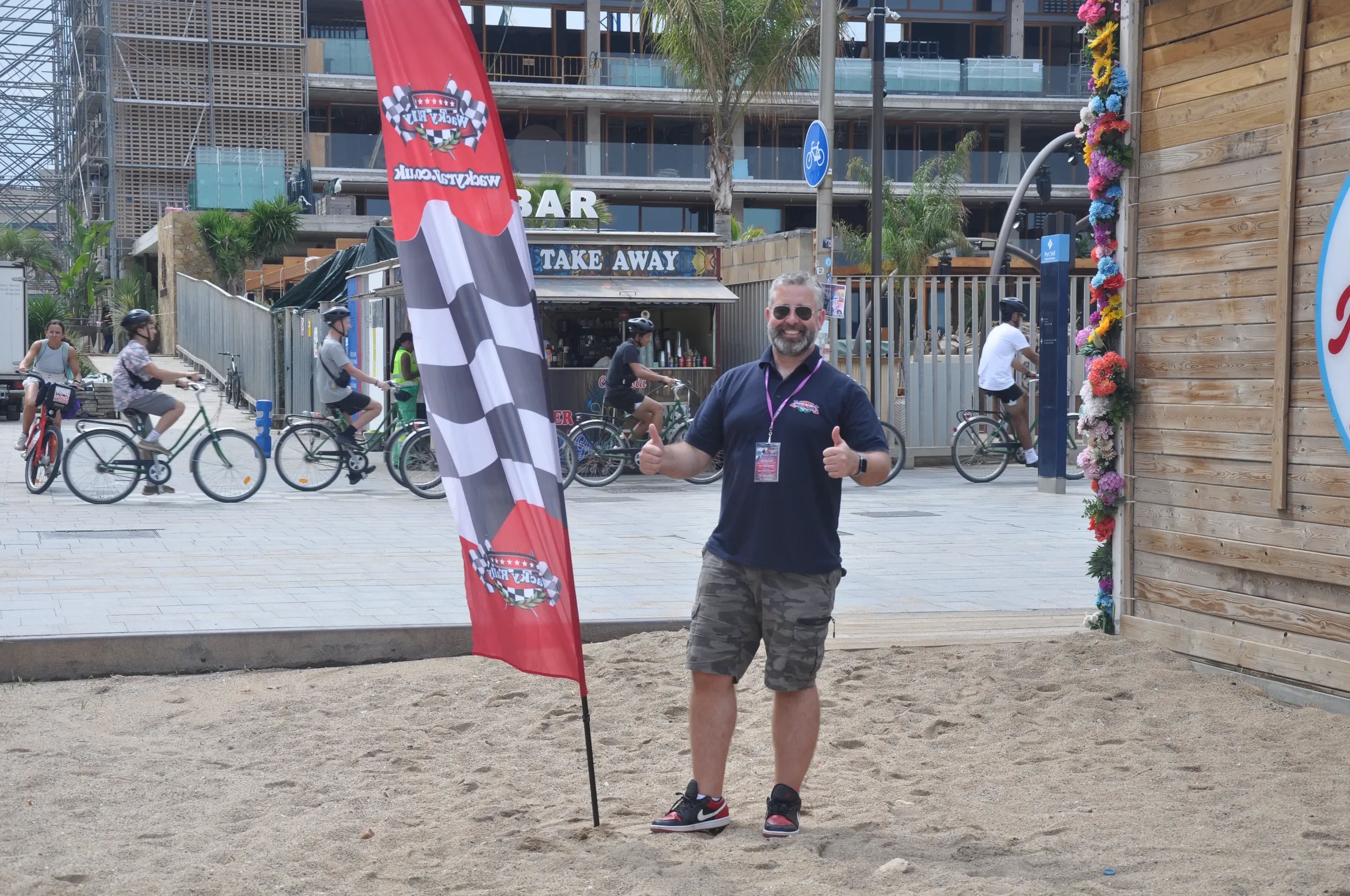 A man smiling and giving two thumbs up while standing next to a race flag on a sandy patch at an outdoor event. People are biking in the background, with a building and a bar and take away stand behind them.