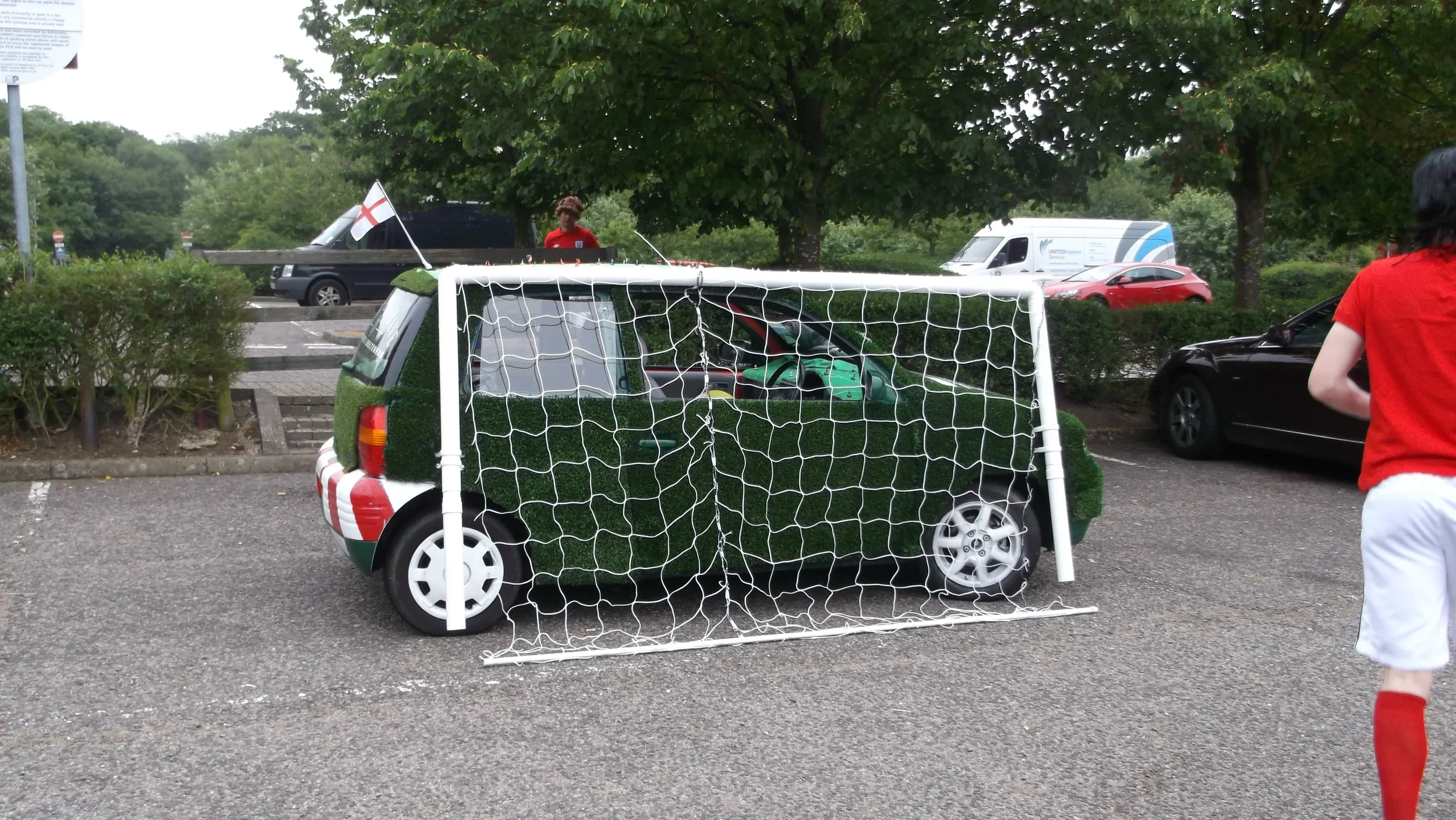 A small green car with artificial grass covering its surface and a soccer goal in front of it, mimicking a soccer scene. The car is parked in a lot surrounded by trees, with a person in a red shirt and white shorts nearby, and several other vehicles 