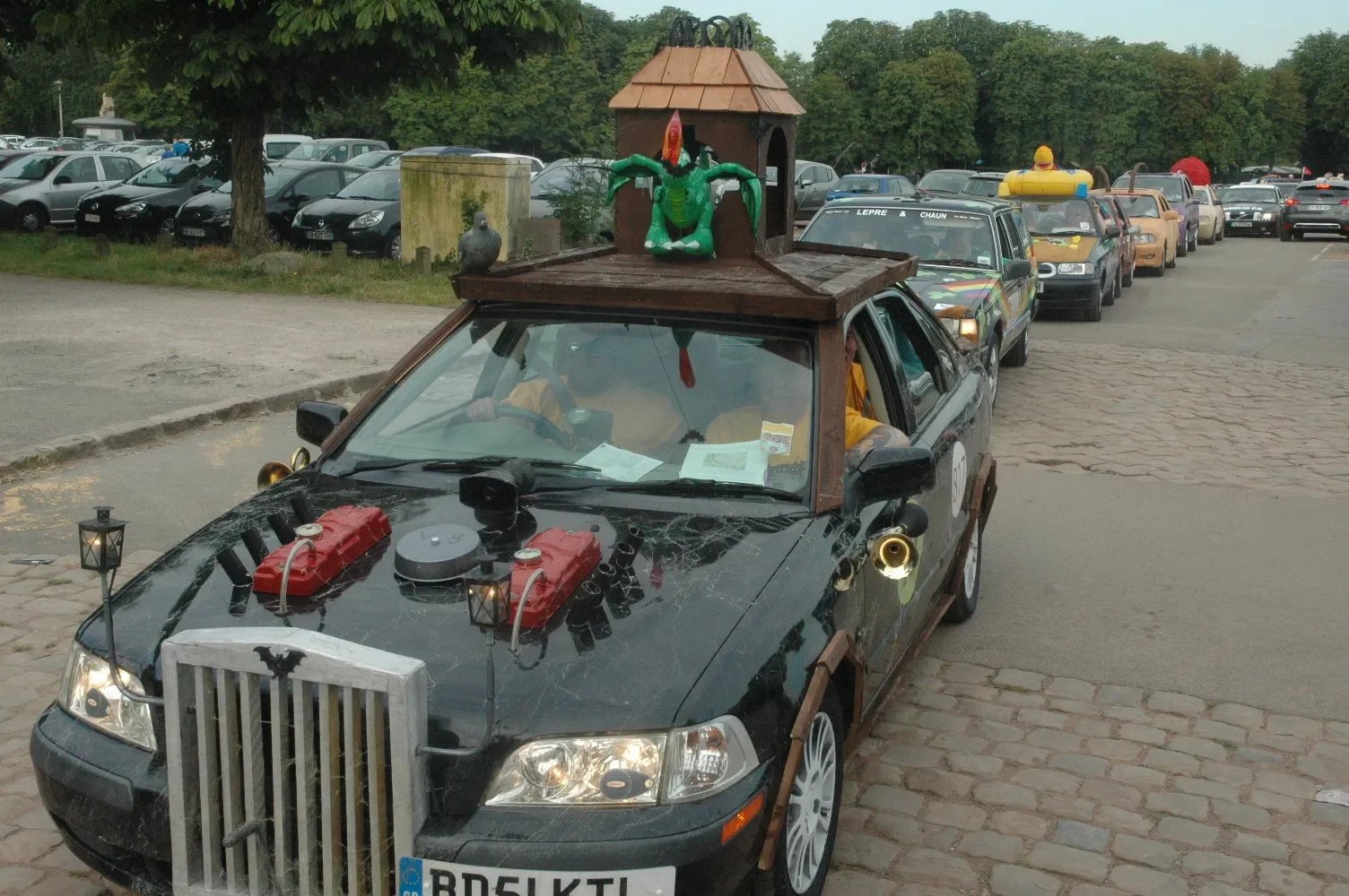 A decorated black car with various objects on and around it, including a small house replica on top, a dragon figure, and lamps. The car is parked on a cobblestone street with a line of similarly decorated cars behind it, and an outdoor parking lot w