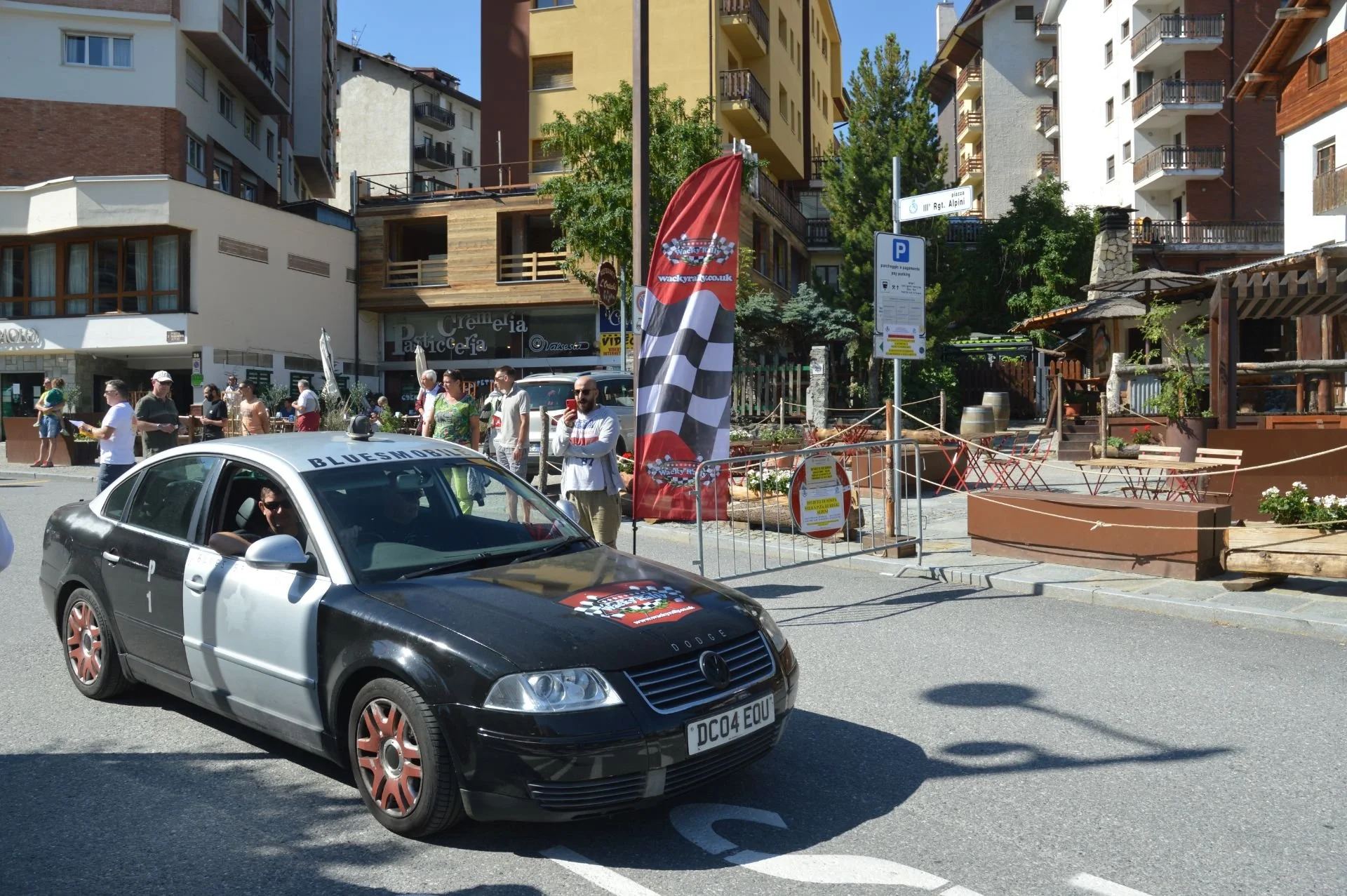 A black and silver car with a ''BLUESHOP'' banner on the windshield driving past an outdoor gathering on a city street. Several people are standing on the sidewalk, some taking pictures. There is a large red and checkered flag and orange barricades s