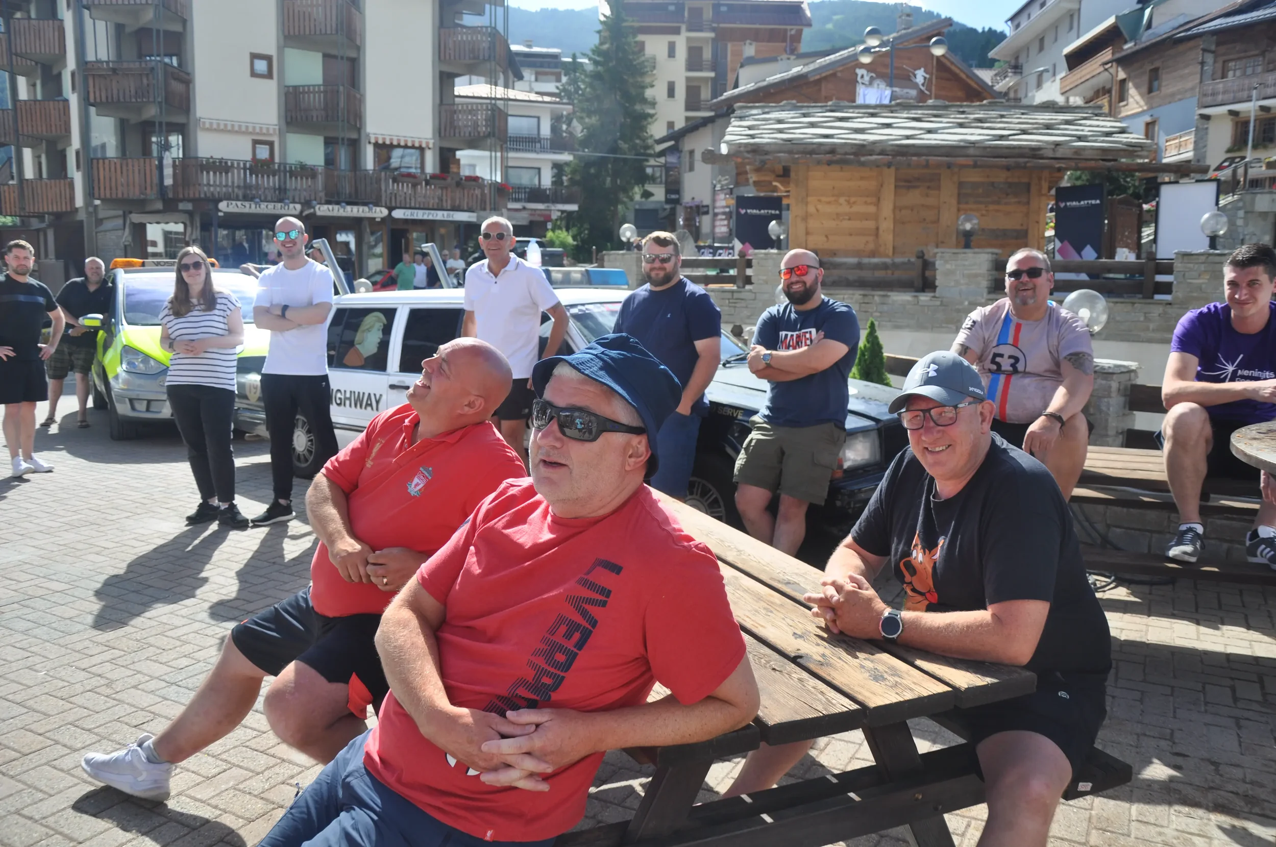 Group of people gathered outdoors in a sunny setting with buildings and mountains in the background, some sitting on benches and some standing, engaging in a social event.