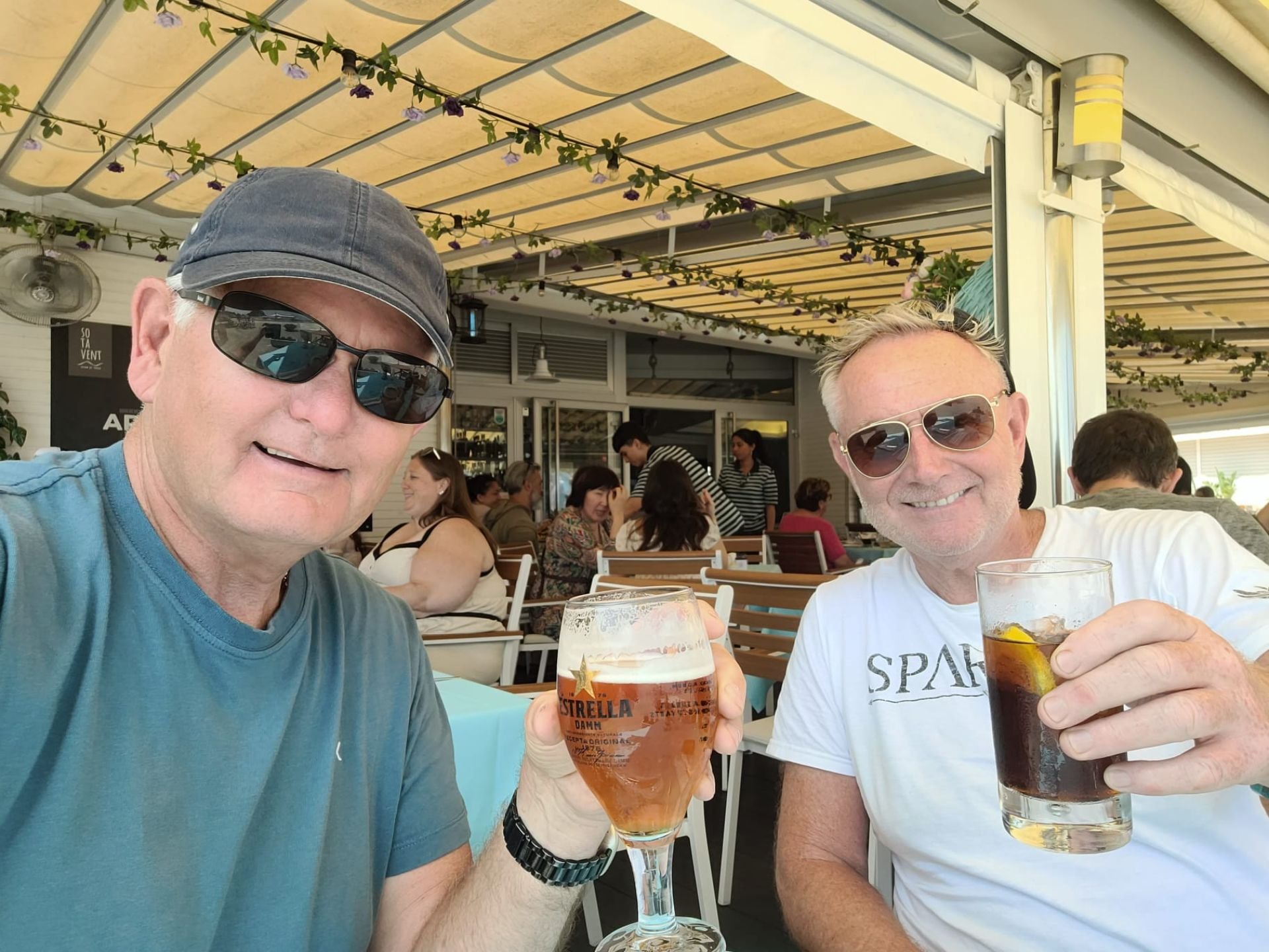 Two men wearing sunglasses sitting at a table in an outdoor restaurant, holding drinks, surrounded by other diners under a canopy decorated with string lights and flowers.